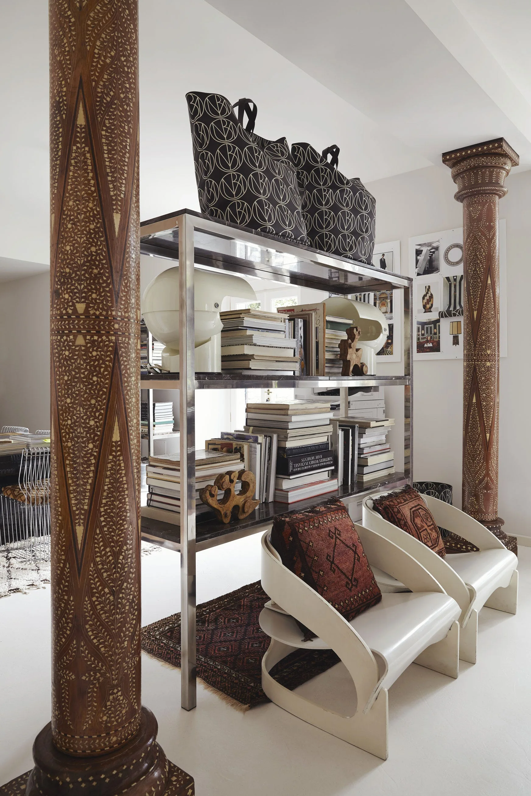 An open metal shelving unit stacked with books is flanked by two intricately carved wooden pillars, with two modern white armchairs in front.