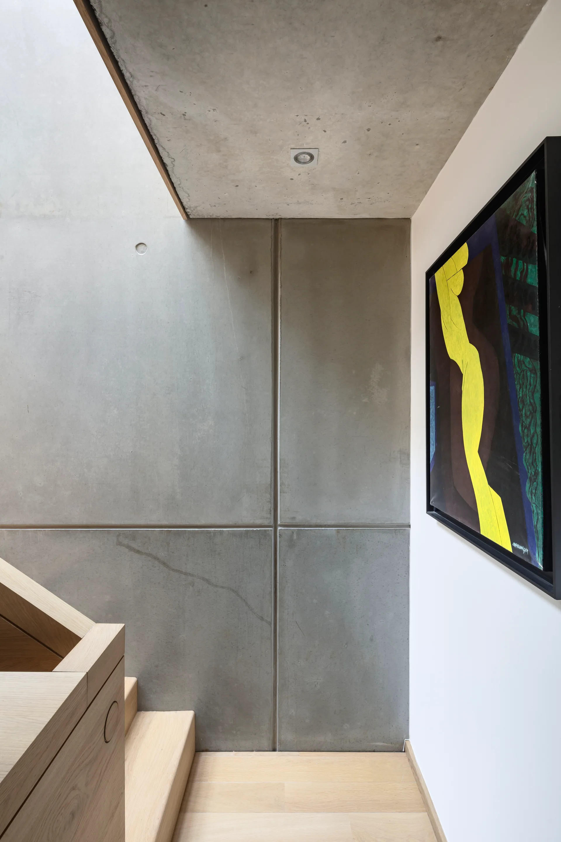 A minimalist staircase featuring light oak wood steps and a handrail alongside industrial grey concrete wall panels and framed colorful artwork.