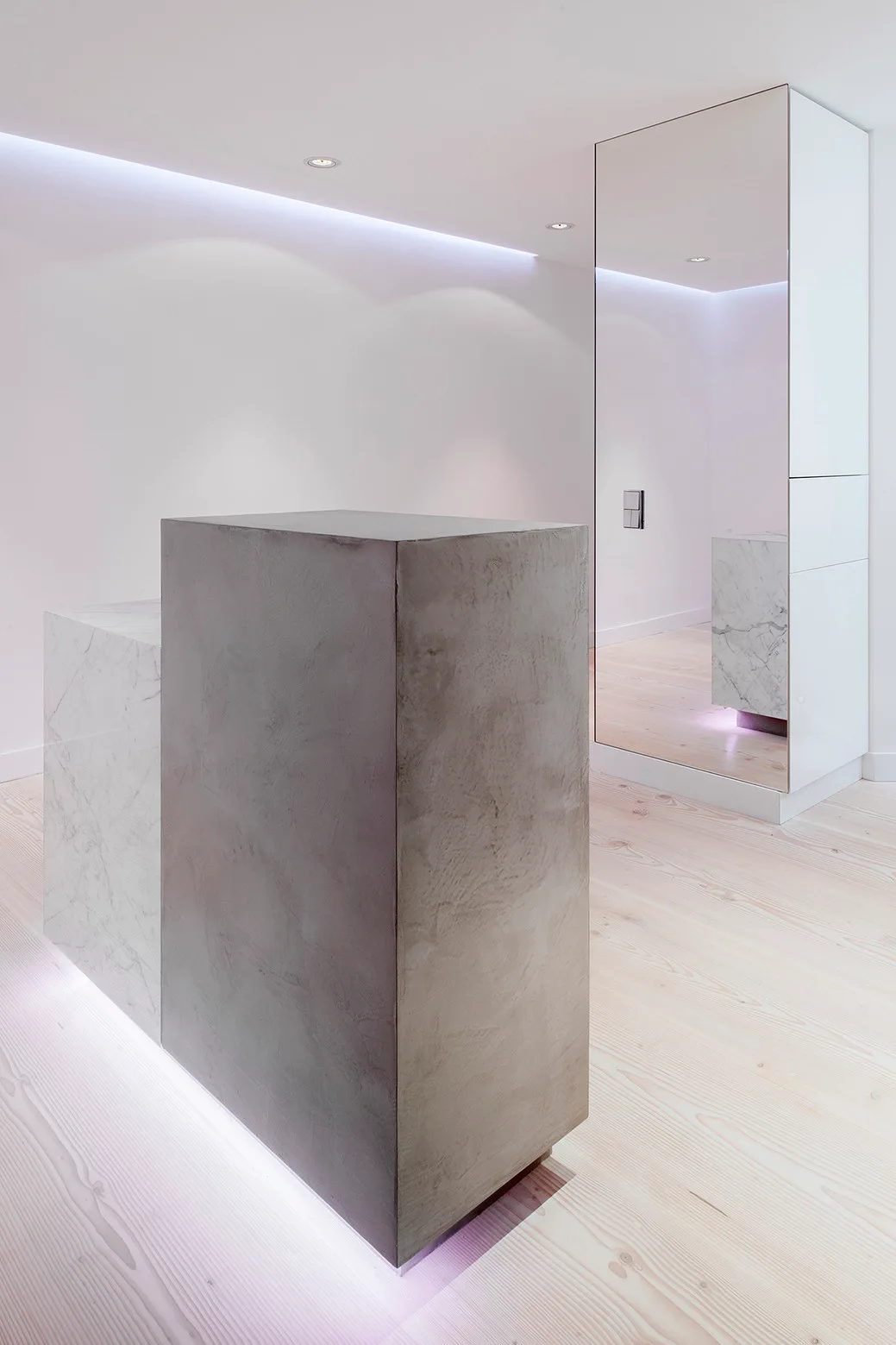 A minimalist reception desk made of marble and textured concrete blocks sits on a pale wood floor beside a tall mirrored wall.