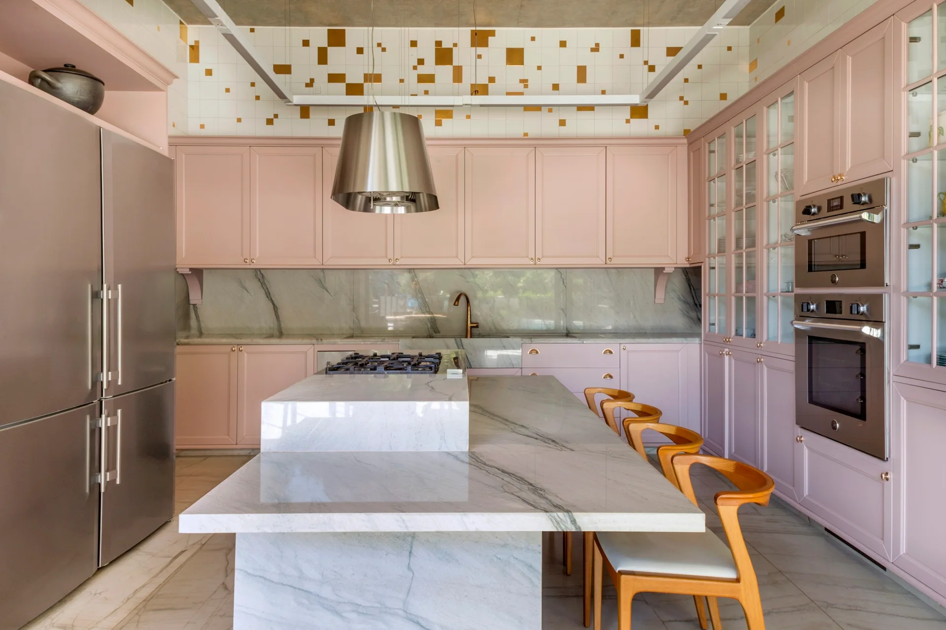 A modern kitchen featuring matte pink cabinets, a tiered veined marble island with wooden stools, and gold-accented upper wall tiles.