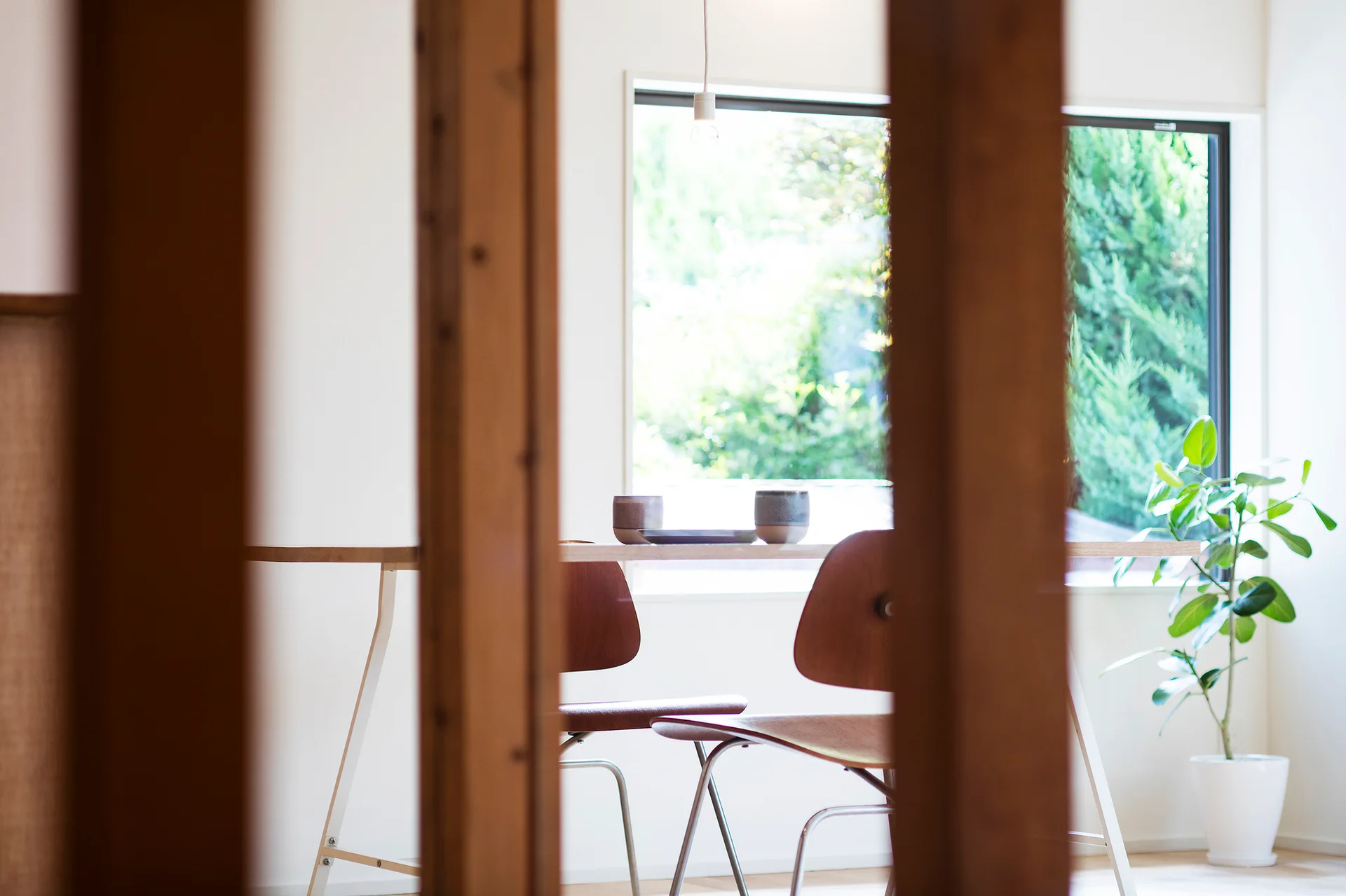 A bright dining area features a light wood table, two mid-century modern chairs, a potted plant, and a large window overlooking a garden.