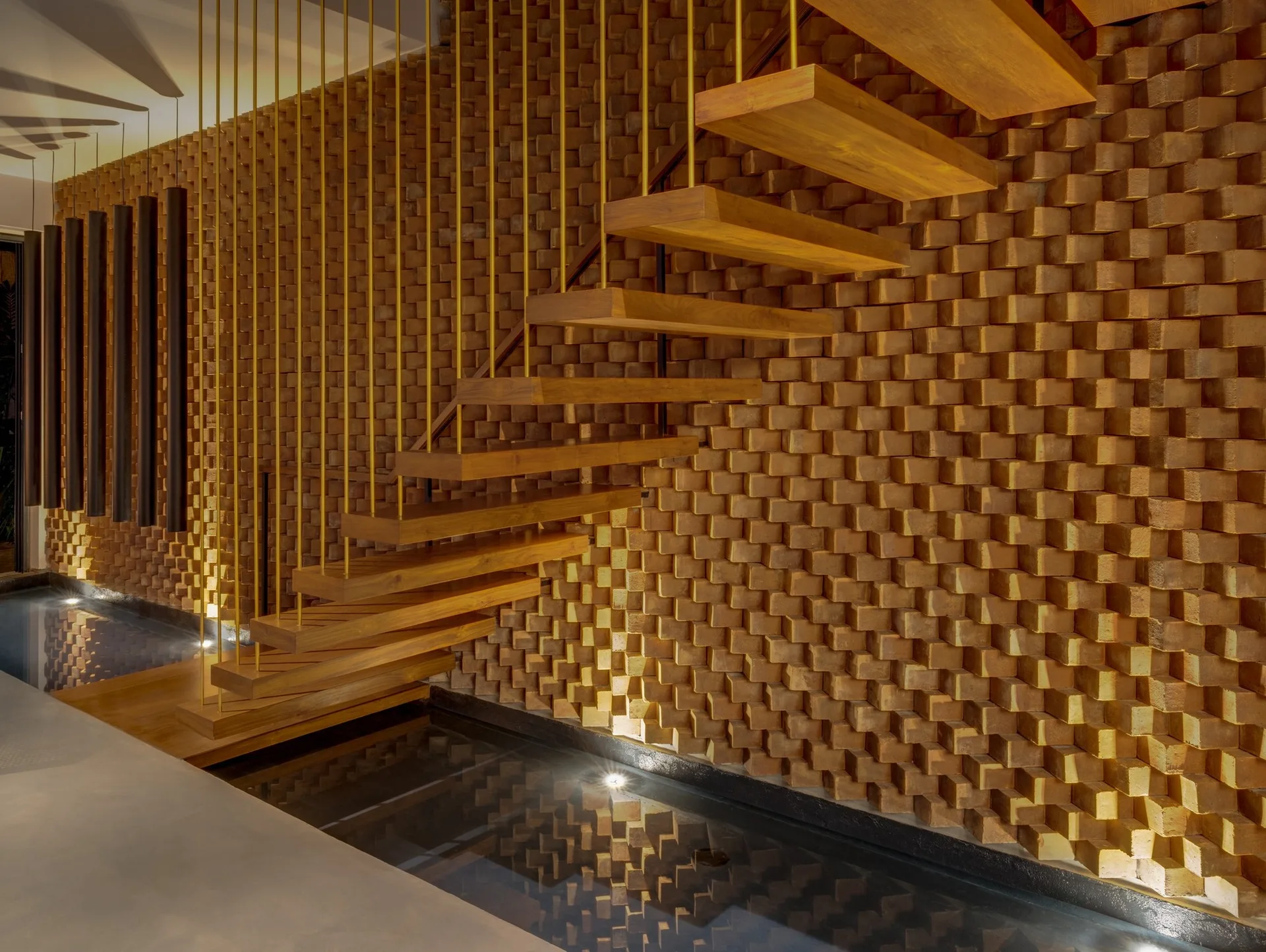 A modern wooden floating staircase features vertical gold metal rod railings set against a three-dimensional brick wall above an indoor reflection pool.