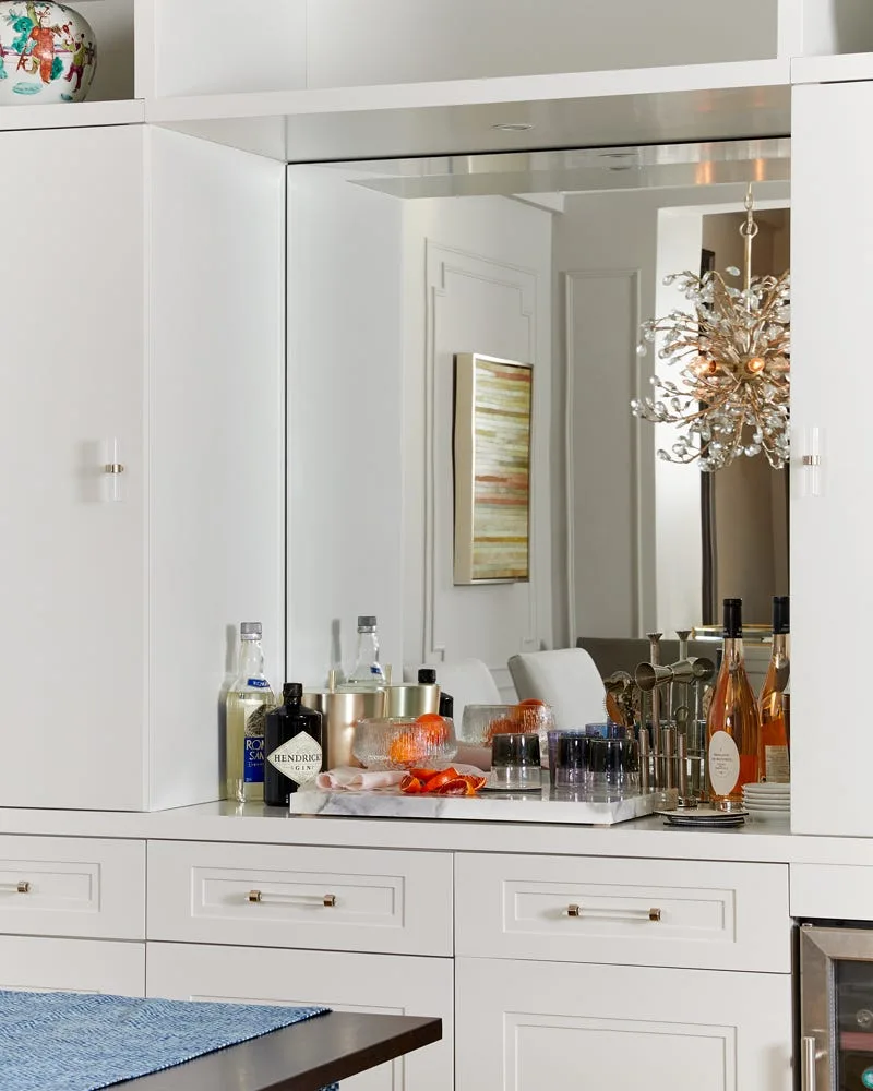 White built-in home bar cabinetry features a mirrored backsplash reflecting a crystal chandelier, with glassware and liquor bottles on a marble tray.