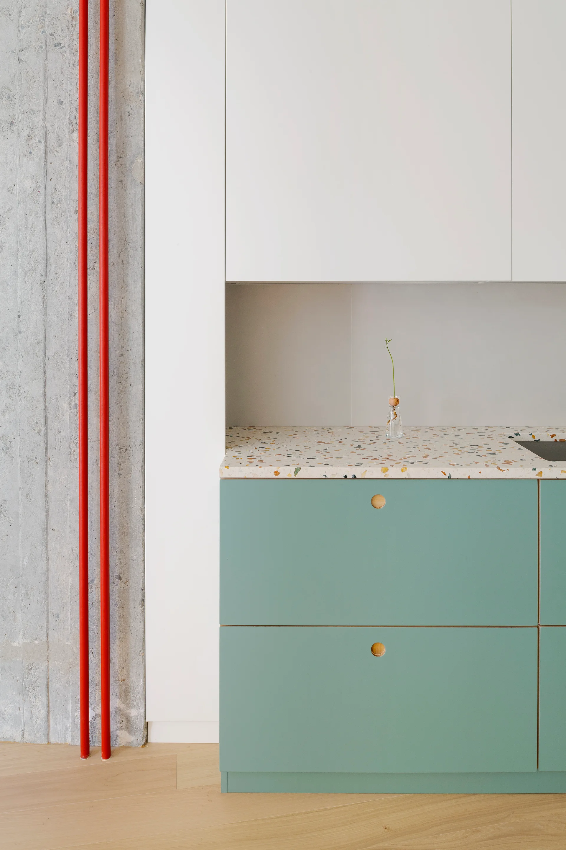 Modern kitchen detail with teal lower cabinets, a speckled terrazzo countertop, white upper cabinetry, and red industrial pipes on a concrete wall.