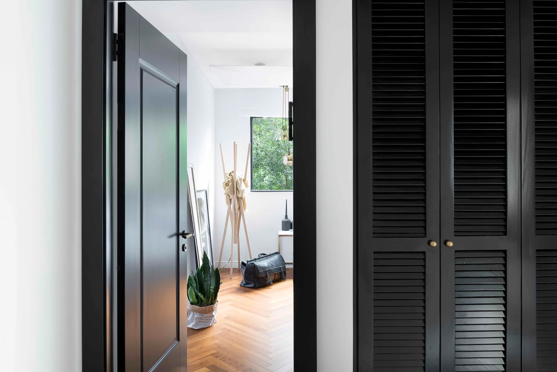 A view through a black doorway reveals an entryway with herringbone wood flooring, black louvered closet doors, and a wooden coat rack.