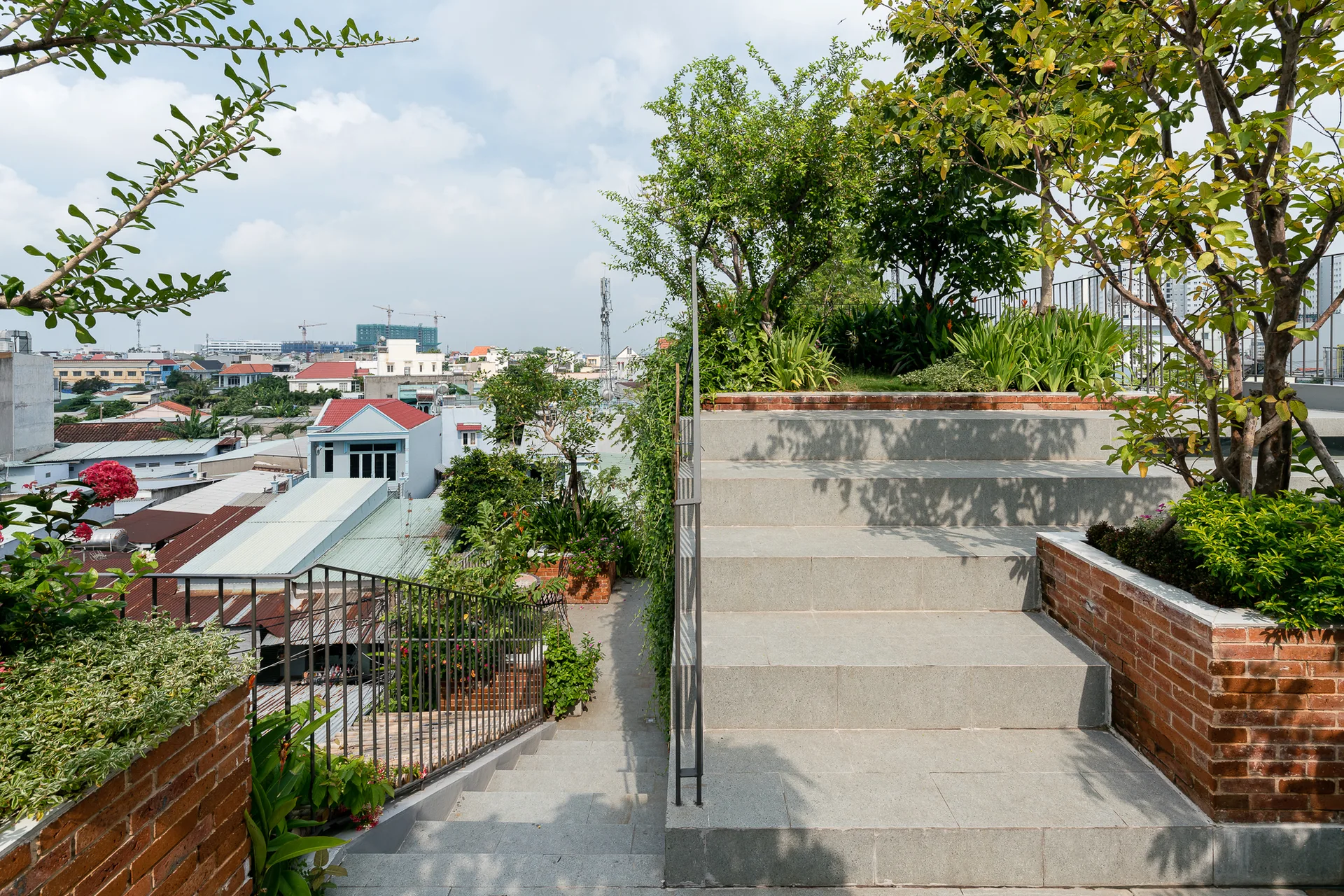An urban rooftop terrace garden features wide stone stairs flanked by red brick planters filled with lush green trees and plants under a blue sky.