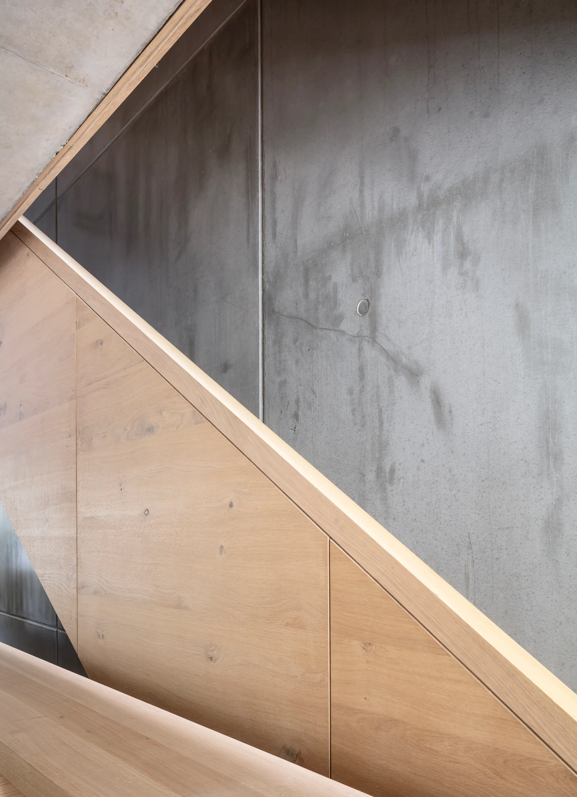 A close-up view of a minimalist staircase featuring light brown wood panels and a railing set against a textured grey concrete wall.