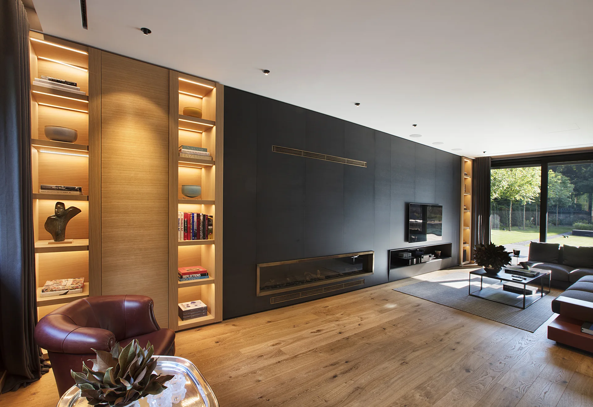 A modern living room features a matte black fireplace wall next to illuminated wood built-in bookshelves and a red leather armchair.