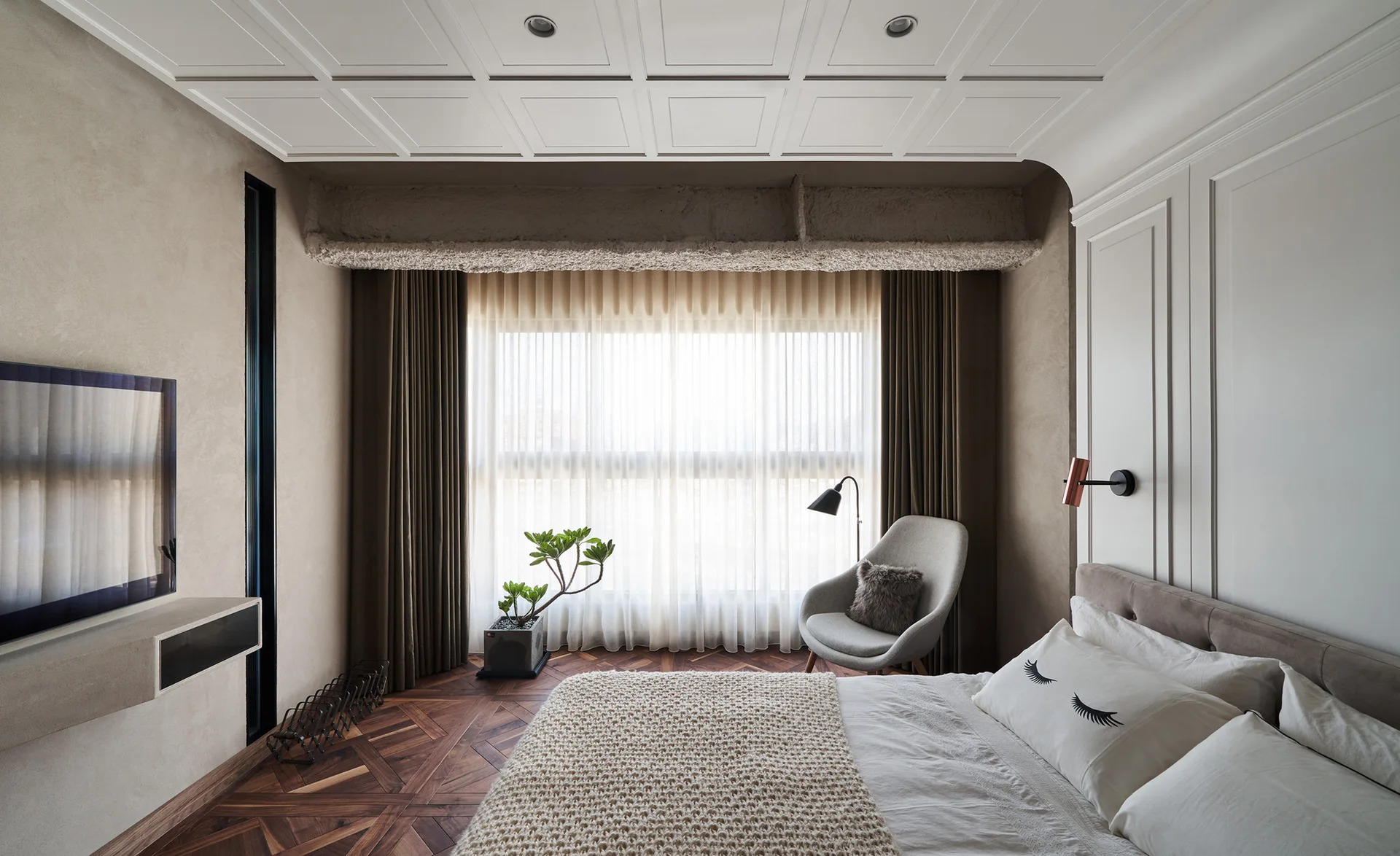 A modern bedroom features a white coffered ceiling, an exposed raw concrete beam above a large window, dark parquet wood floors, and a bed.