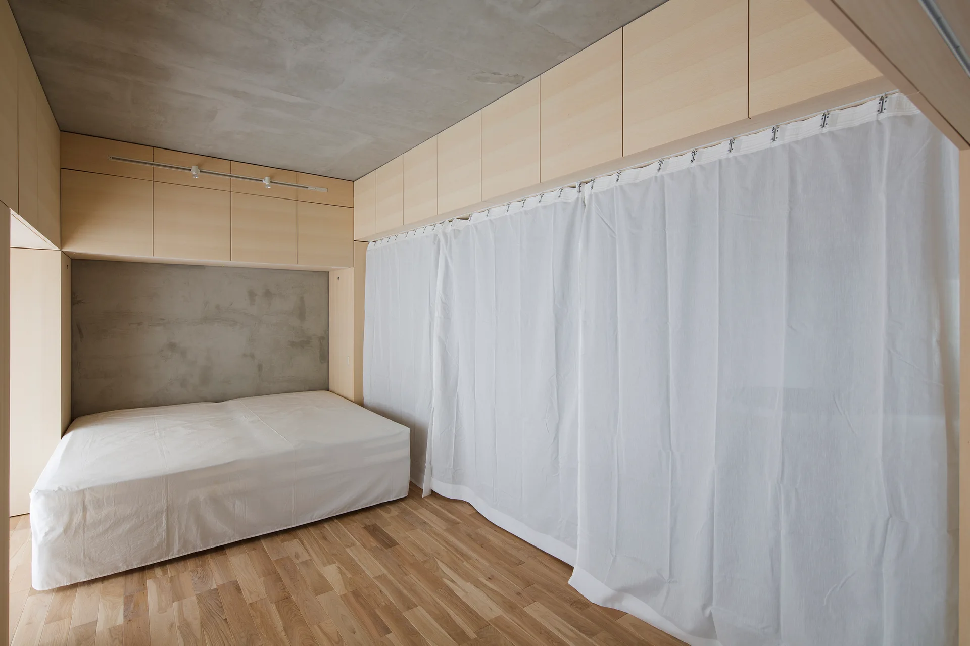 A minimalist bedroom features a low platform bed beneath light wood cabinets, set against a raw concrete ceiling and wall with white curtains.