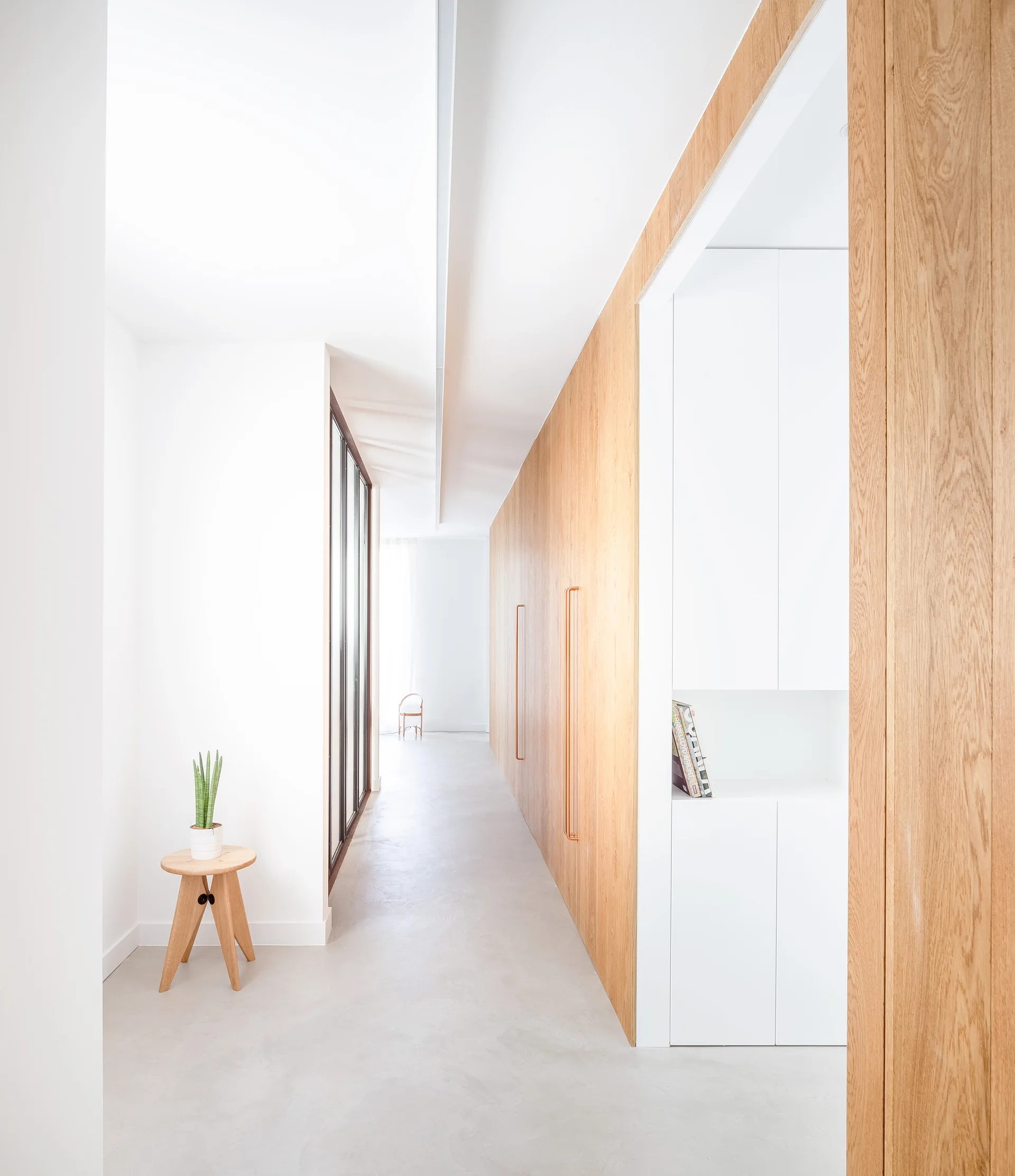A minimalist white hallway features a full-height wall of light oak wood cabinets with long metallic handles and a concrete-style floor.
