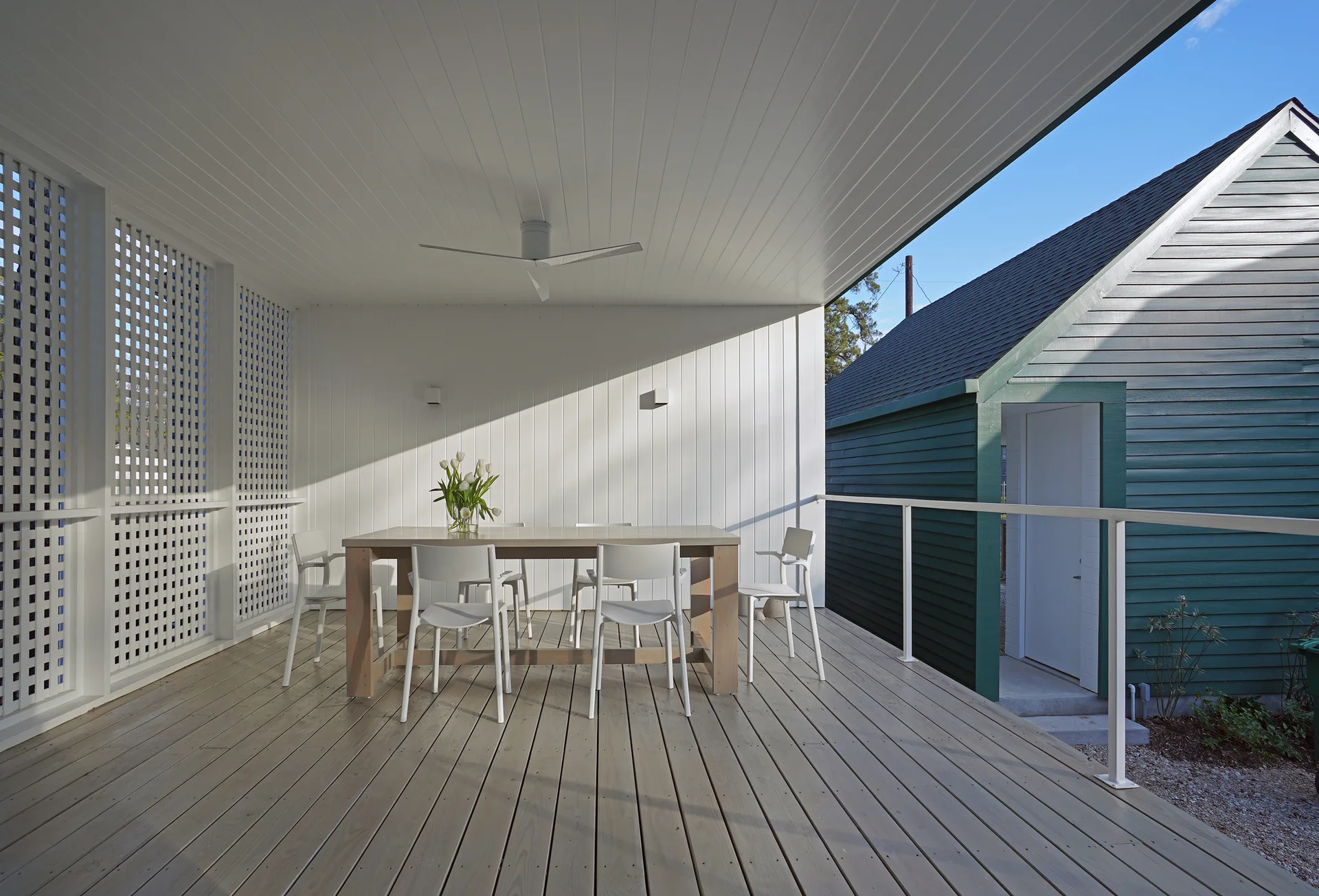 A covered wood deck features a minimalist wood dining table and white chairs set against a white paneled wall and a white lattice screen.