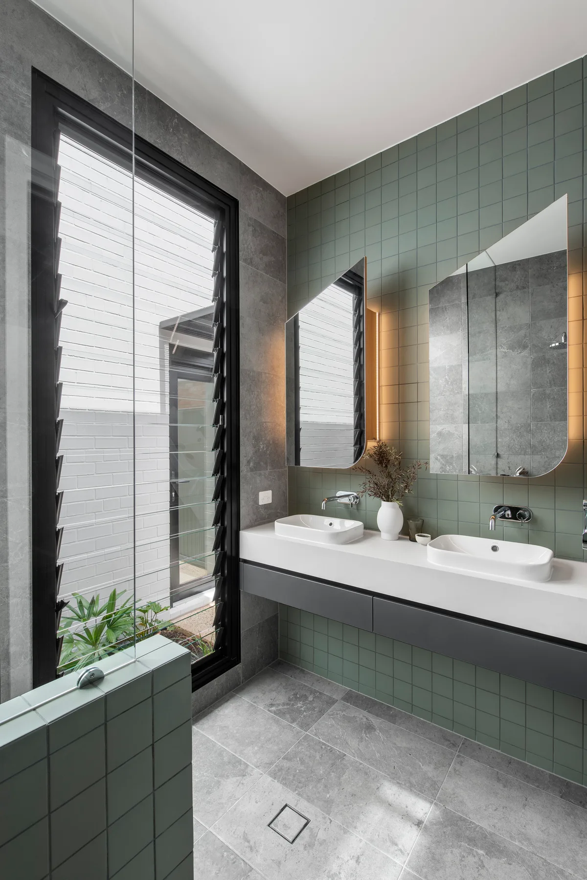 A modern bathroom features a sage green grid tiled wall, a white floating double vanity, and grey stone floor tiles.