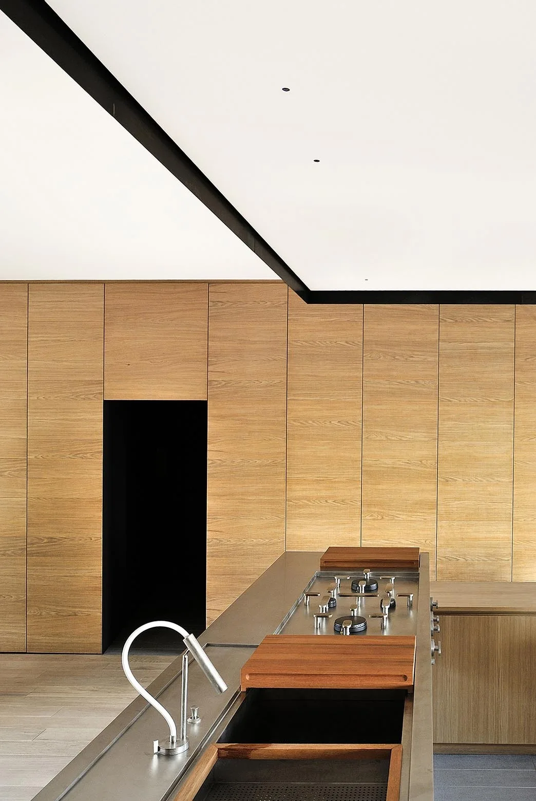 A minimalist kitchen features a stainless steel island with an integrated stove and faucet set against a full wall of wood-paneled cabinetry.