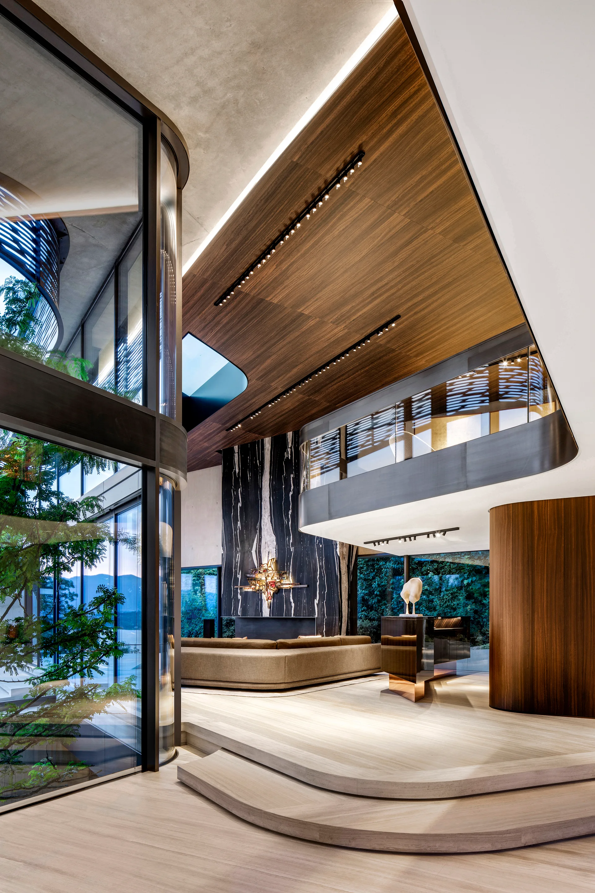 A modern double-height living room features a sunken beige sectional sofa, a black veined marble fireplace wall, a wood ceiling, and curved glass windows.