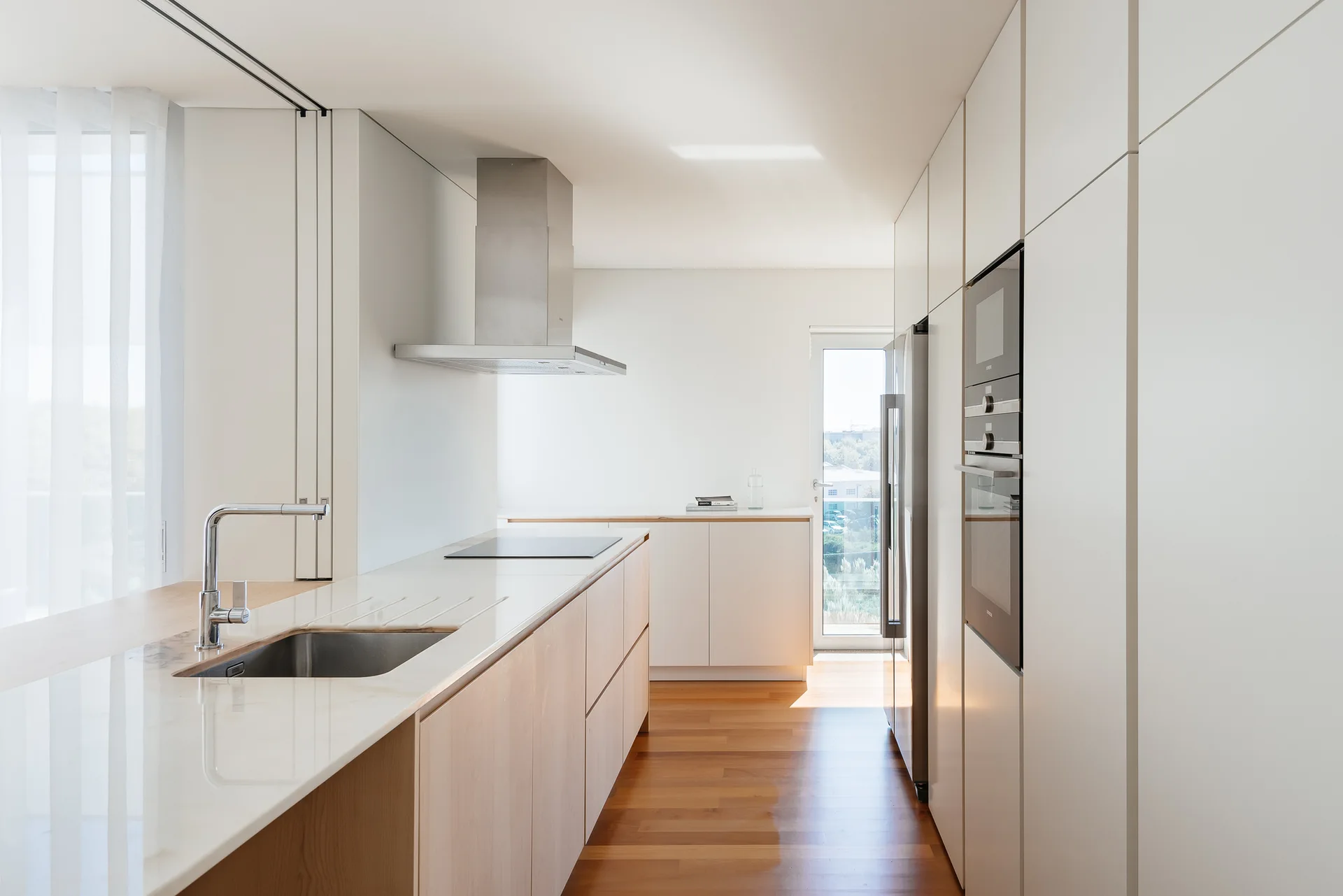 A minimalist galley kitchen featuring handleless white cabinets, a light wood island with a stone countertop, and hardwood flooring.