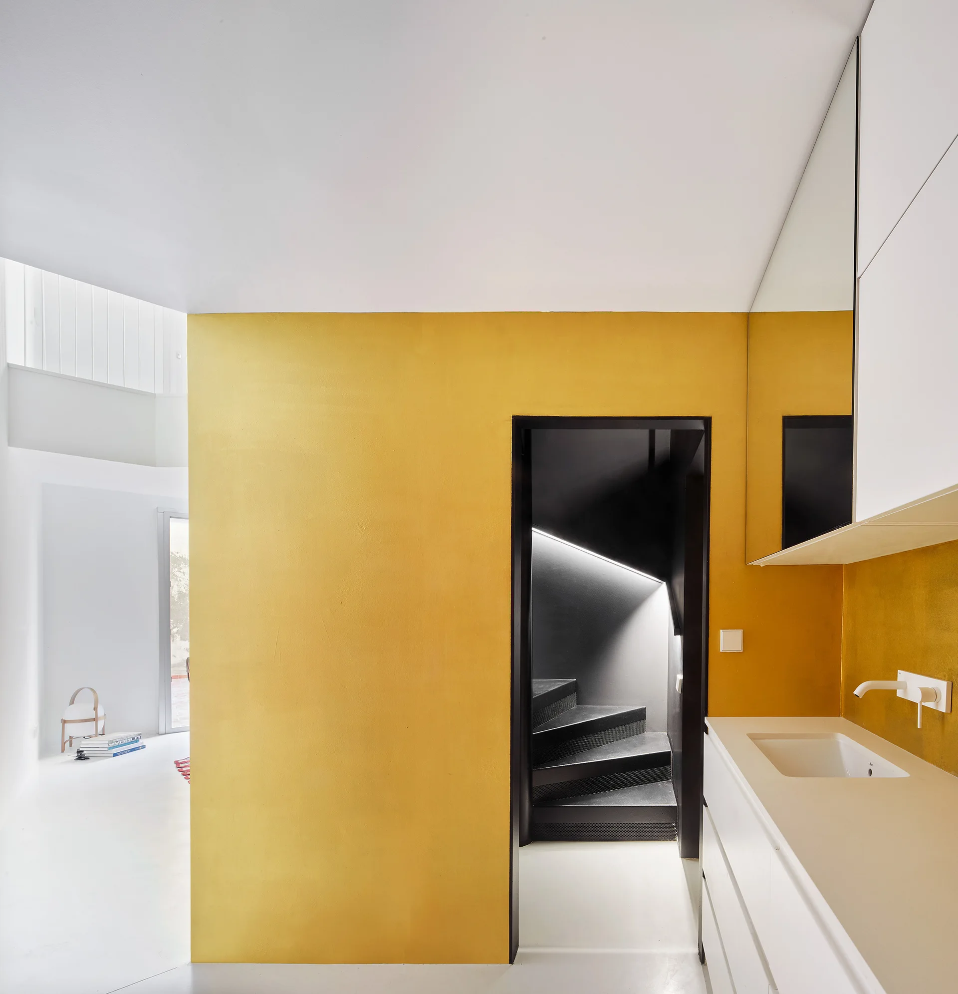 A bright yellow partition wall stands between a minimalist white bathroom vanity and a dark staircase with a linear light strip.