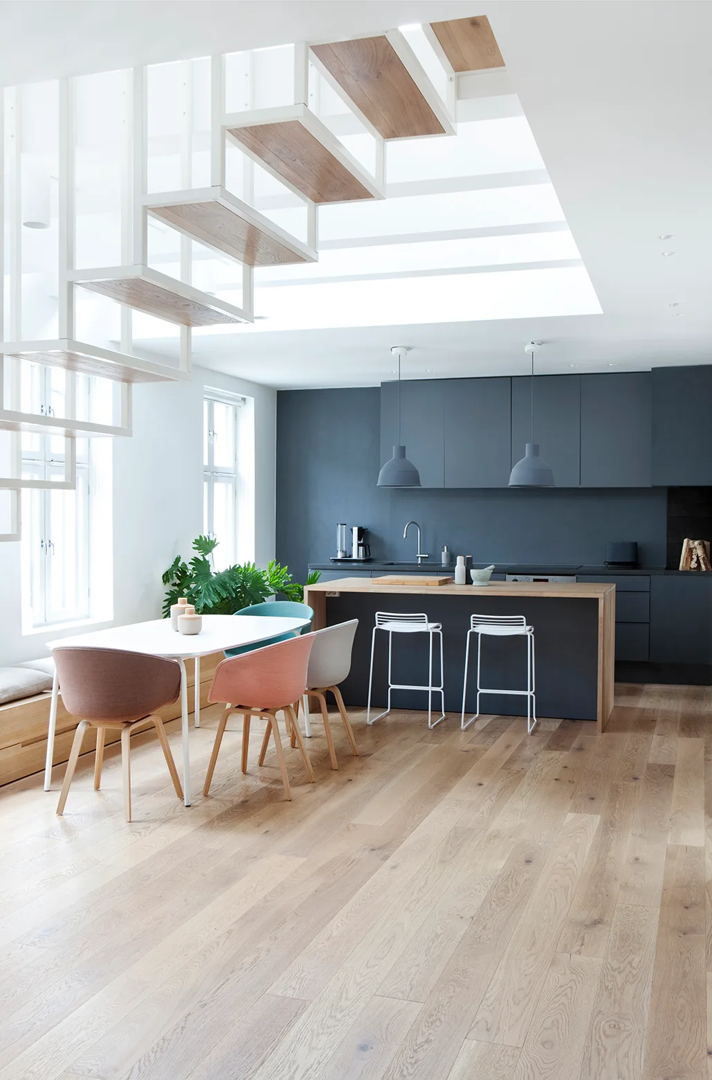 A light-filled modern kitchen displays a dark gray wall of cabinets, a wood-topped island, colorful dining chairs, and a white floating staircase.