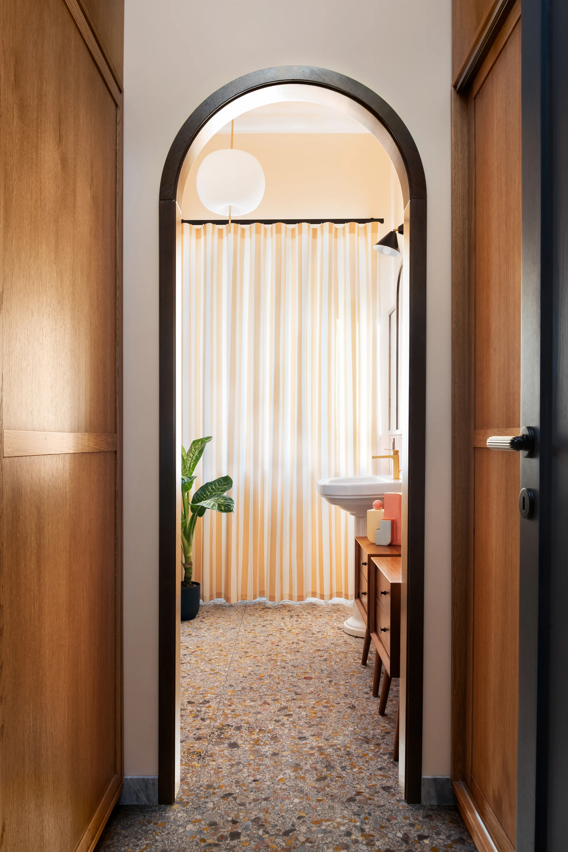 A view through a dark wood arched doorway into a bathroom with terrazzo flooring, a white pedestal sink, and a yellow striped shower curtain.