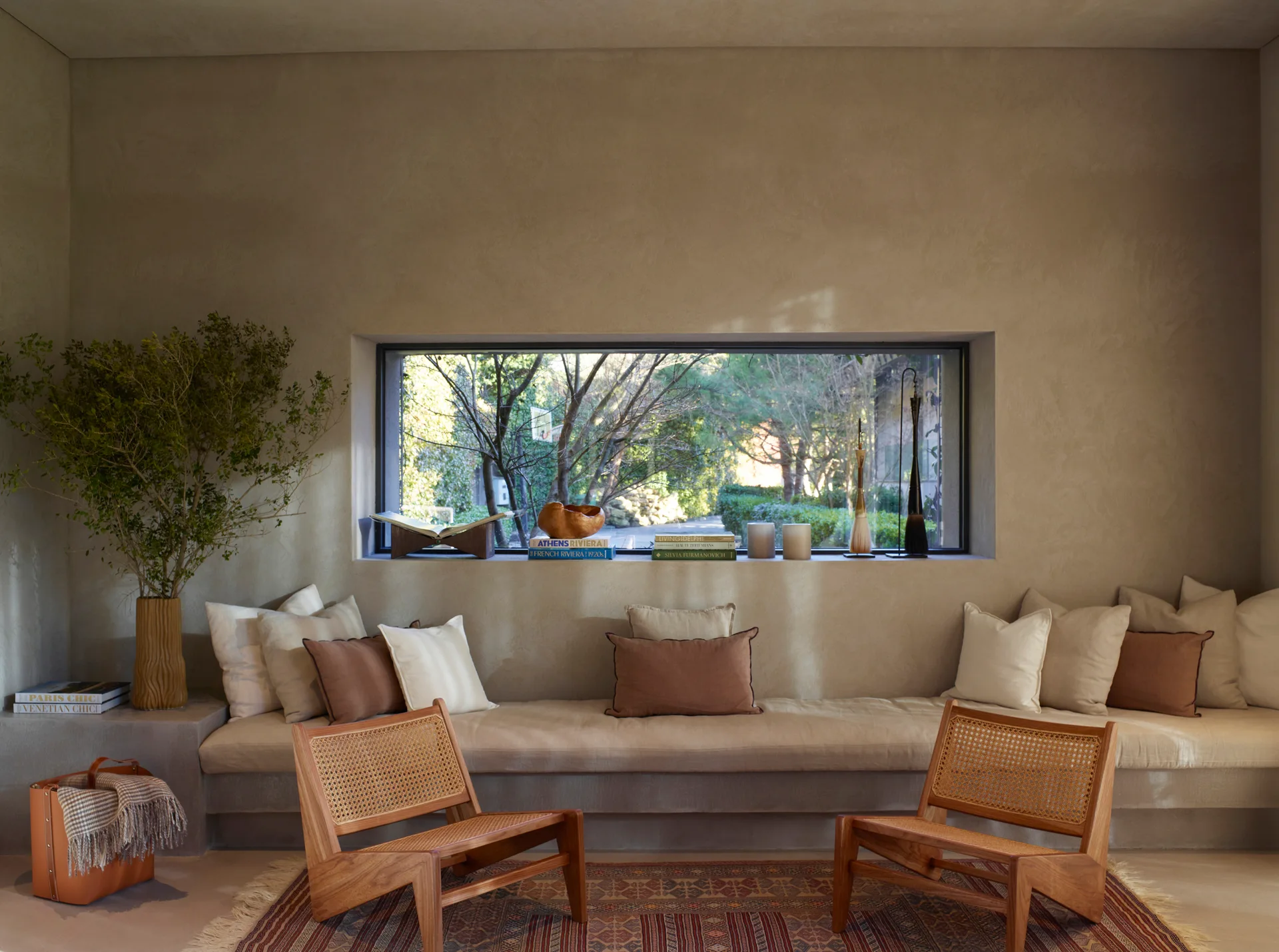 A long built-in plaster bench with linen pillows sits under a large rectangular picture window, paired with two wooden cane armchairs on a patterned rug.