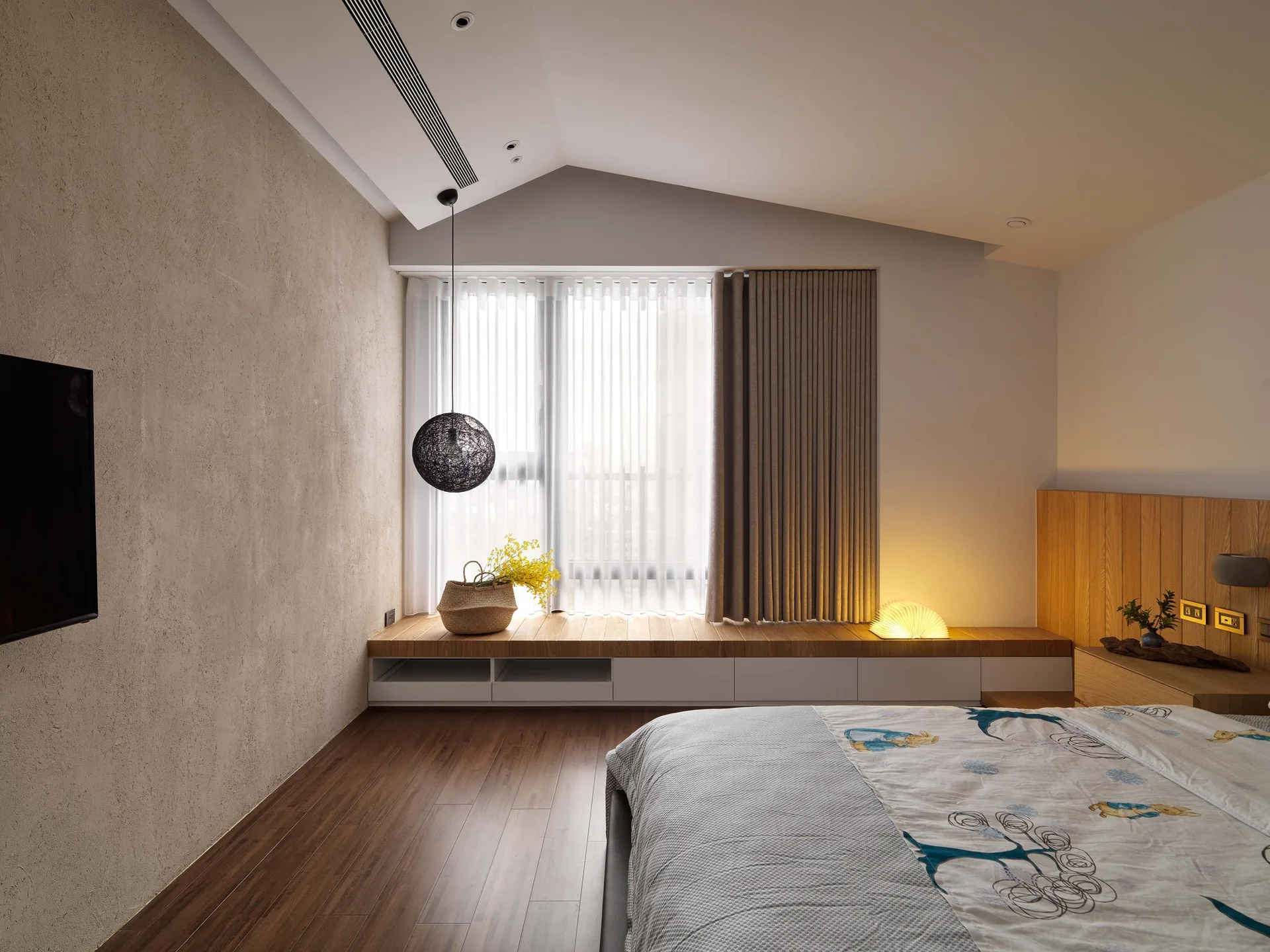 A modern bedroom featuring a built-in wooden window bench, a textured plaster accent wall, a sloped ceiling, and a low platform bed.
