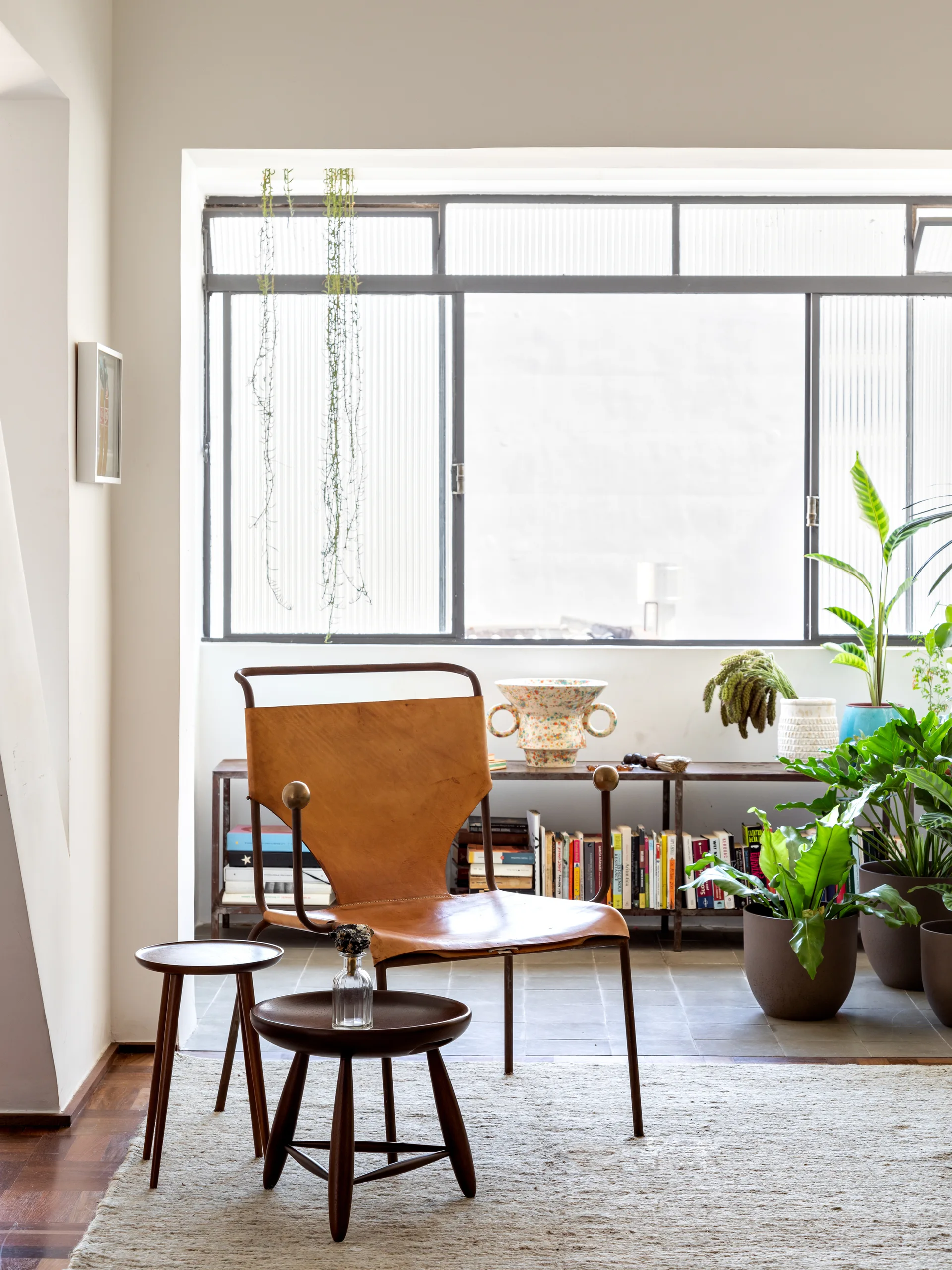 A bright room features a tan leather armchair on a textured beige rug beside two small wooden side tables in front of a large metal-framed window.
