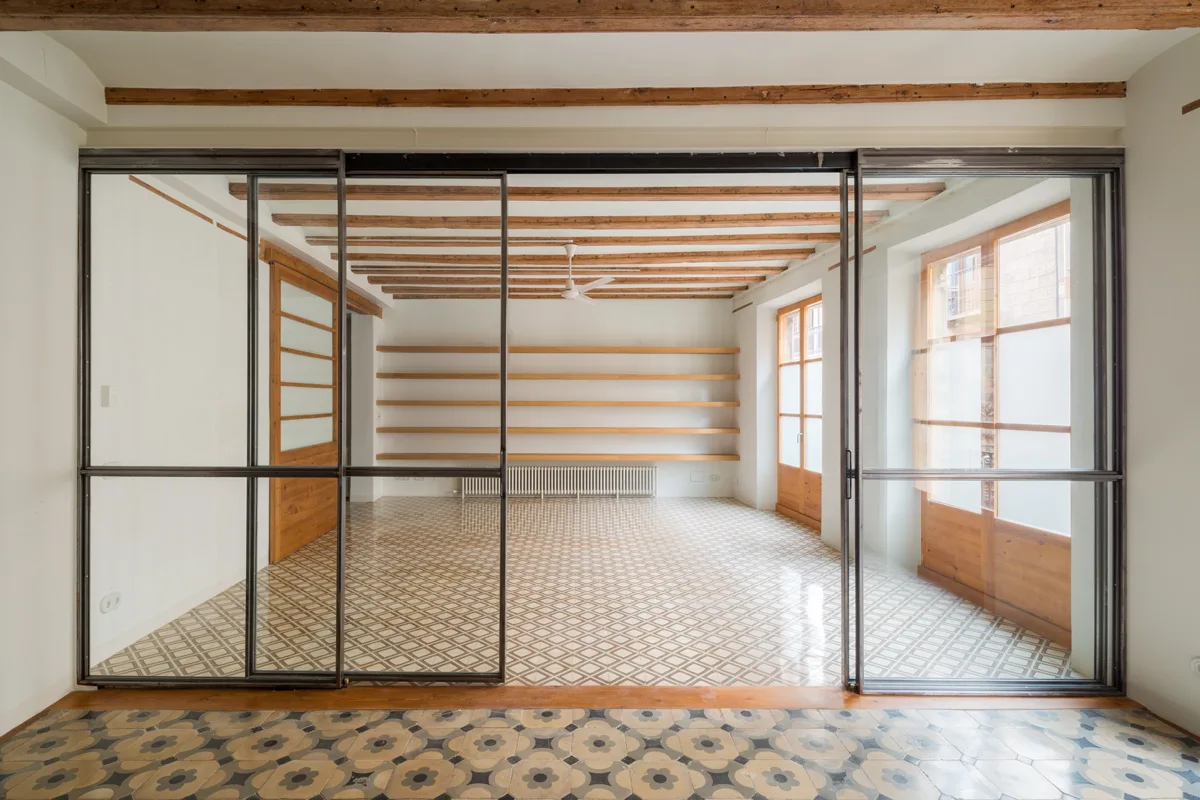 A large black-framed sliding glass partition divides an empty room featuring geometric floor tiles, exposed wood ceiling beams, and wall shelving.