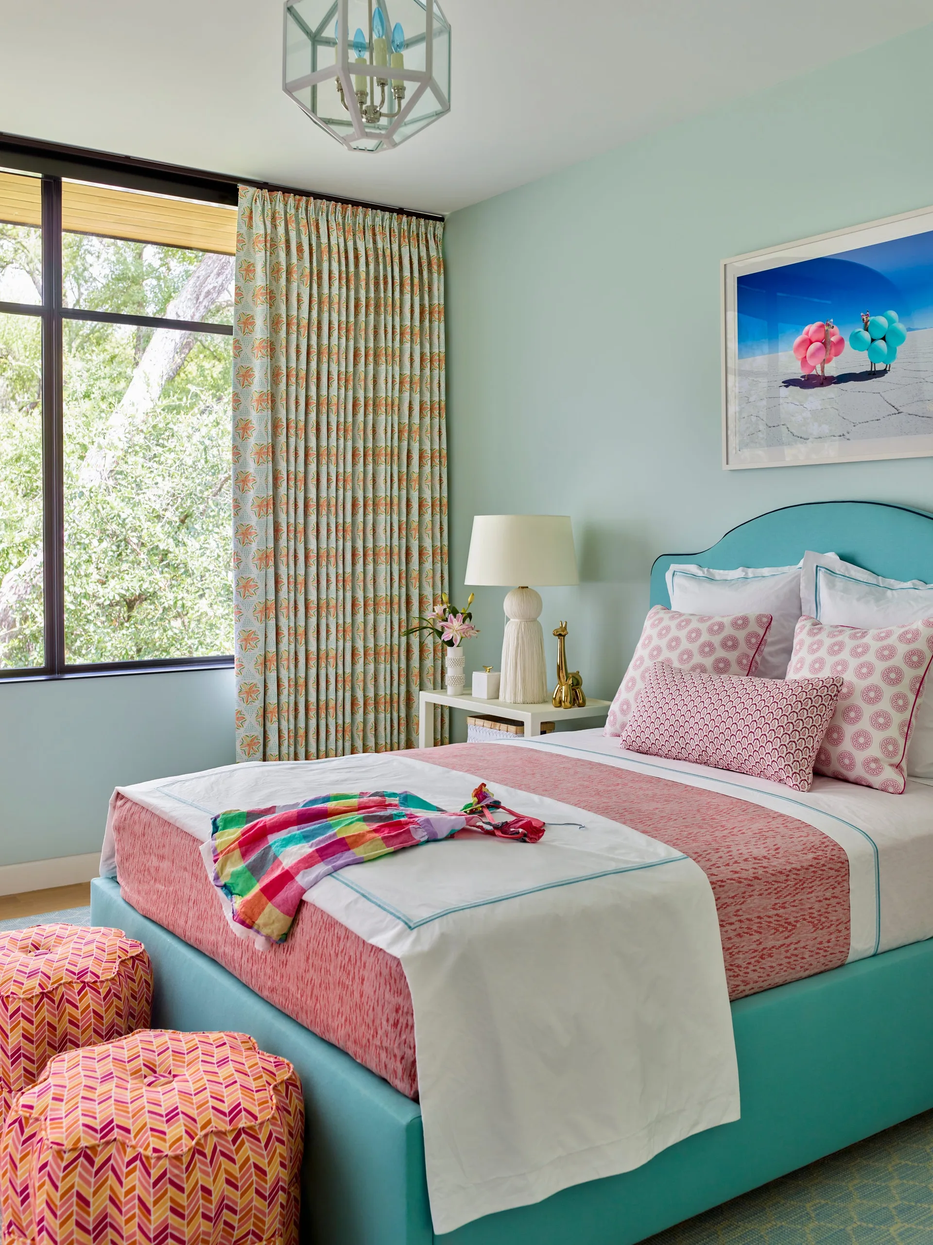 A light blue bedroom features a turquoise upholstered headboard, pink patterned pillows, a white table lamp, and patterned curtains by a large window.