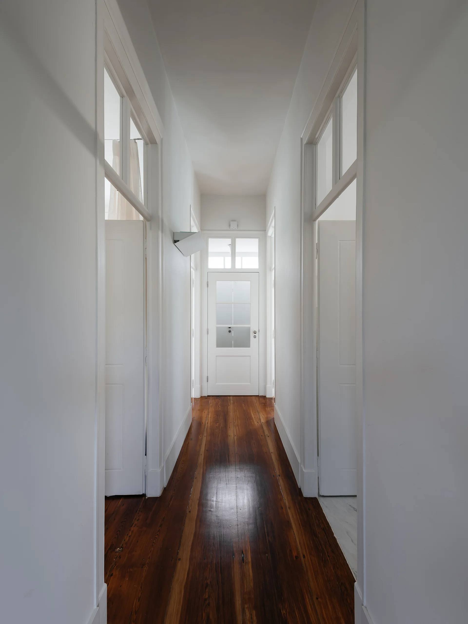 A long white residential hallway featuring dark wood plank flooring, multiple paneled doors, and glass transom windows that let in natural light.