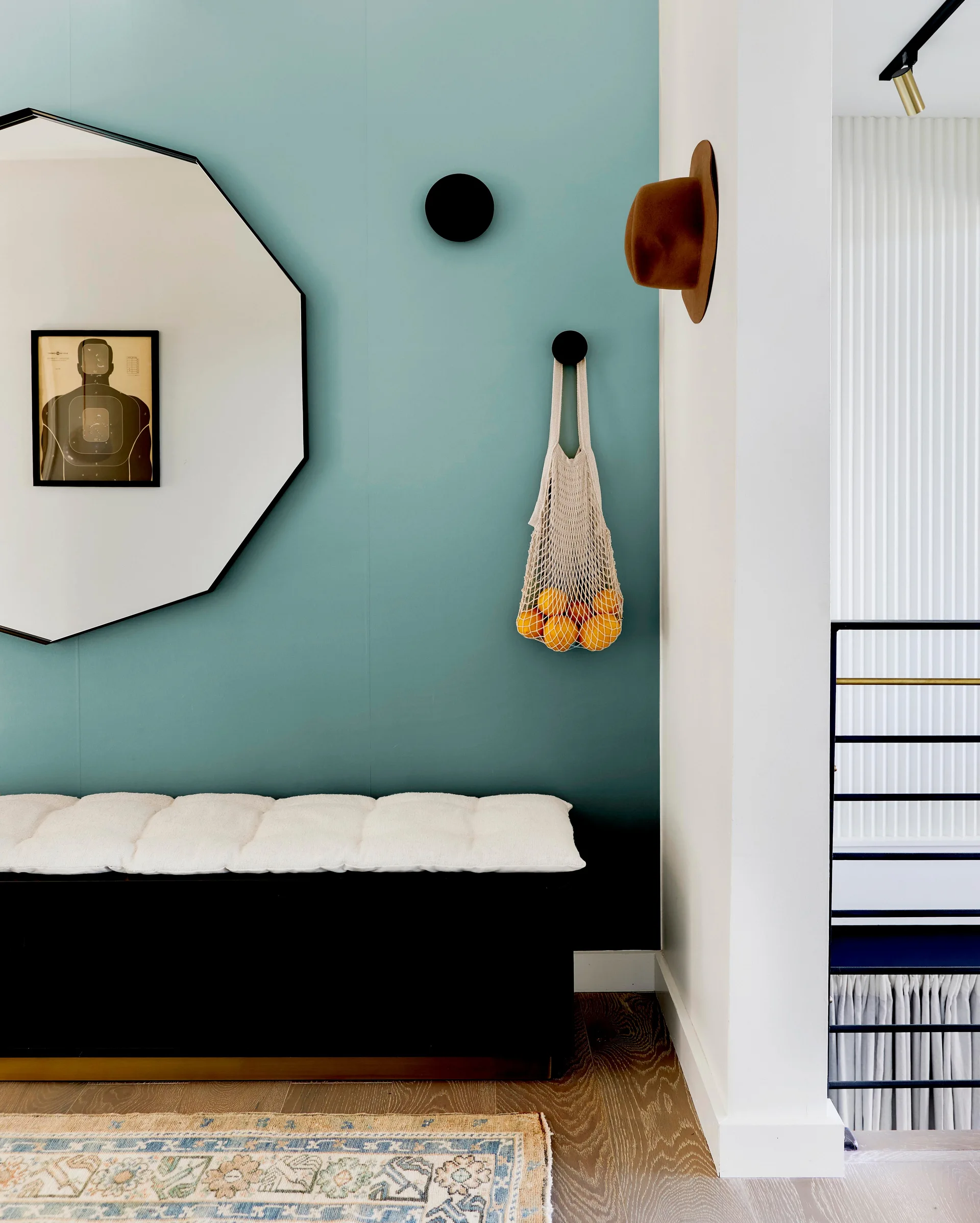 A modern entryway features a black cushioned bench set against a teal accent wall with a large black-framed octagonal mirror and round wall hooks.