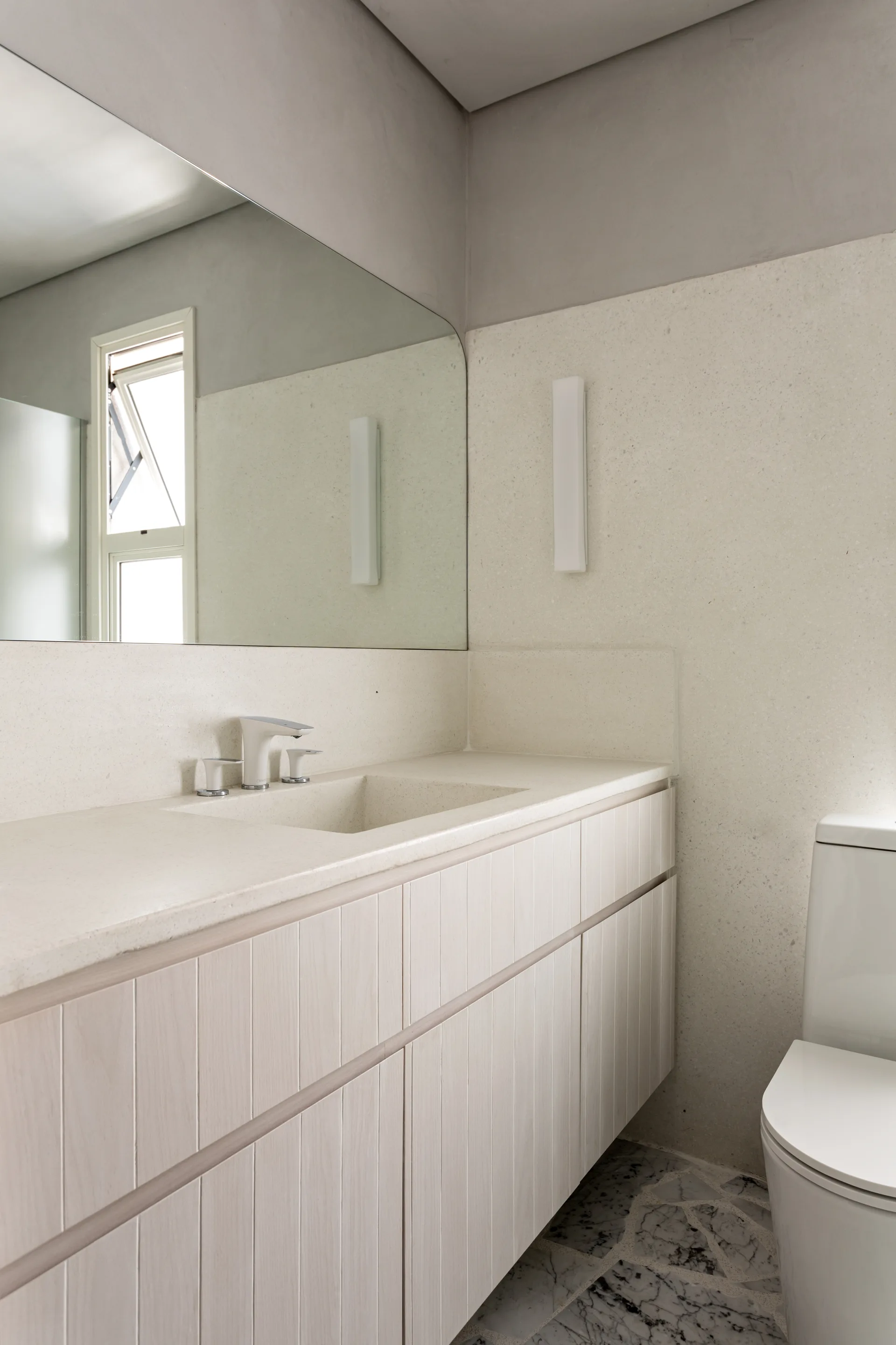 A modern bathroom vanity featuring a light-toned slatted wood cabinet, integrated cream stone countertop, and large frameless mirror.