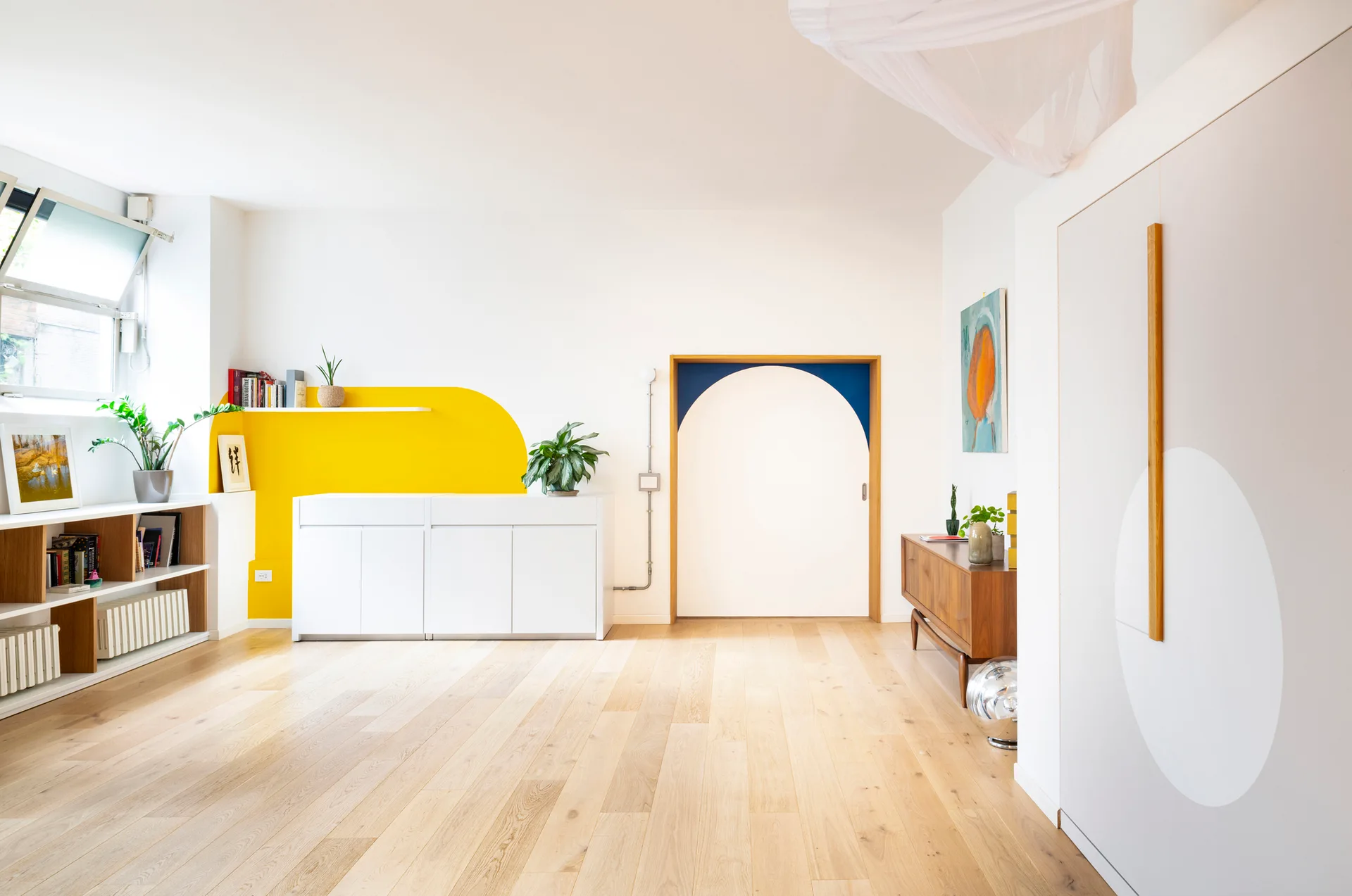 A bright living room with light hardwood floors, a white cabinet against a yellow wall graphic, and a blue painted archway.