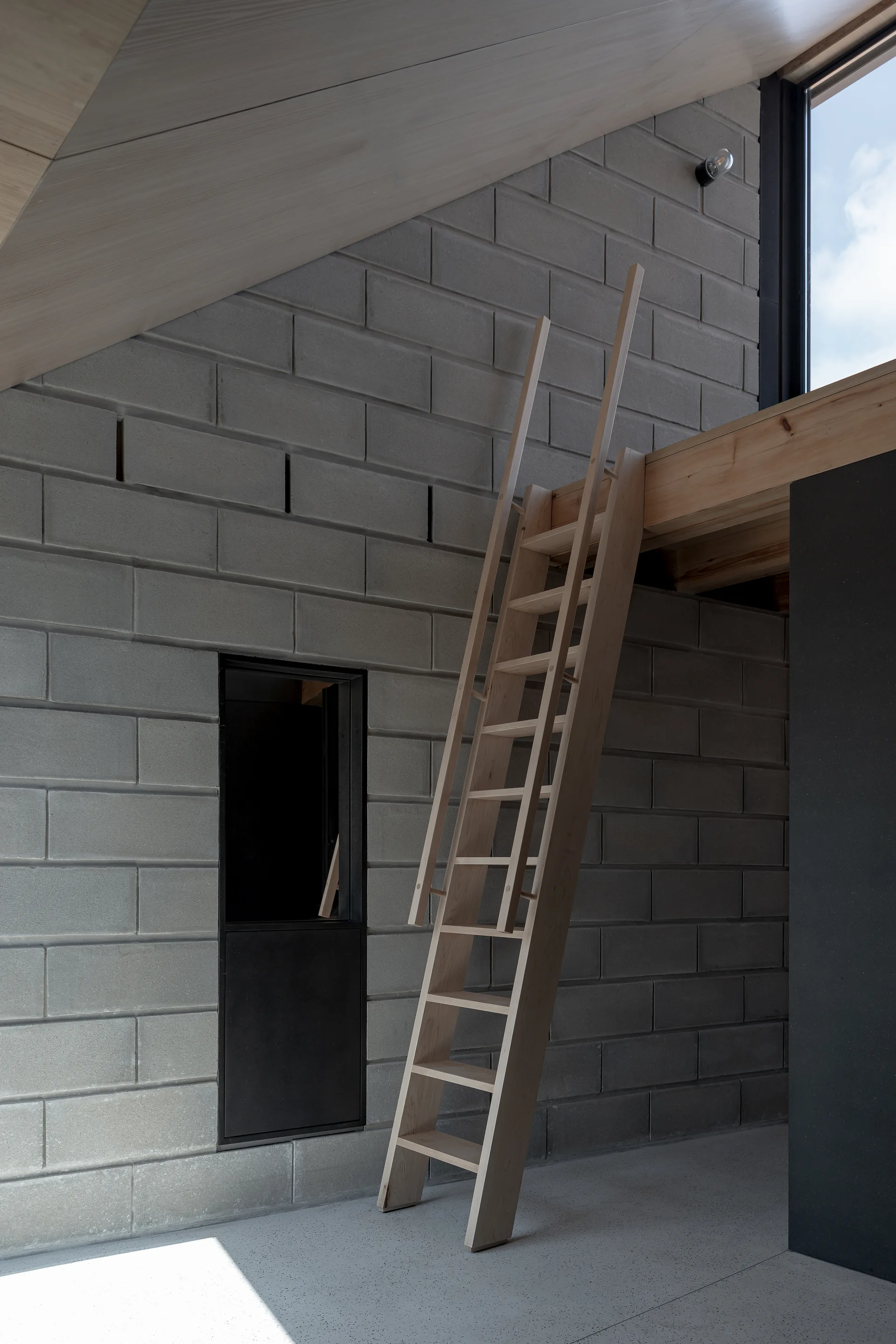 A wooden ladder leans against a light timber loft platform next to an exposed concrete block wall under a sloped wooden ceiling.