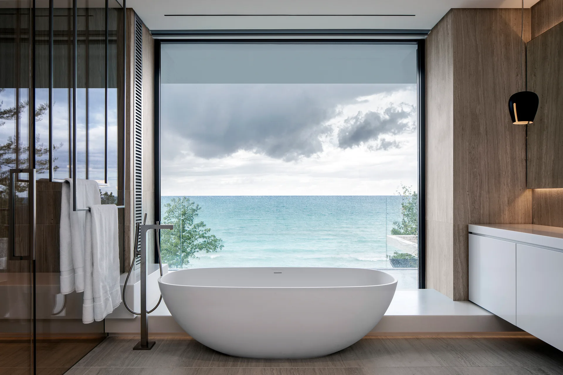A white freestanding bathtub sits in front of a large picture window overlooking the ocean in a contemporary stone-tiled bathroom.