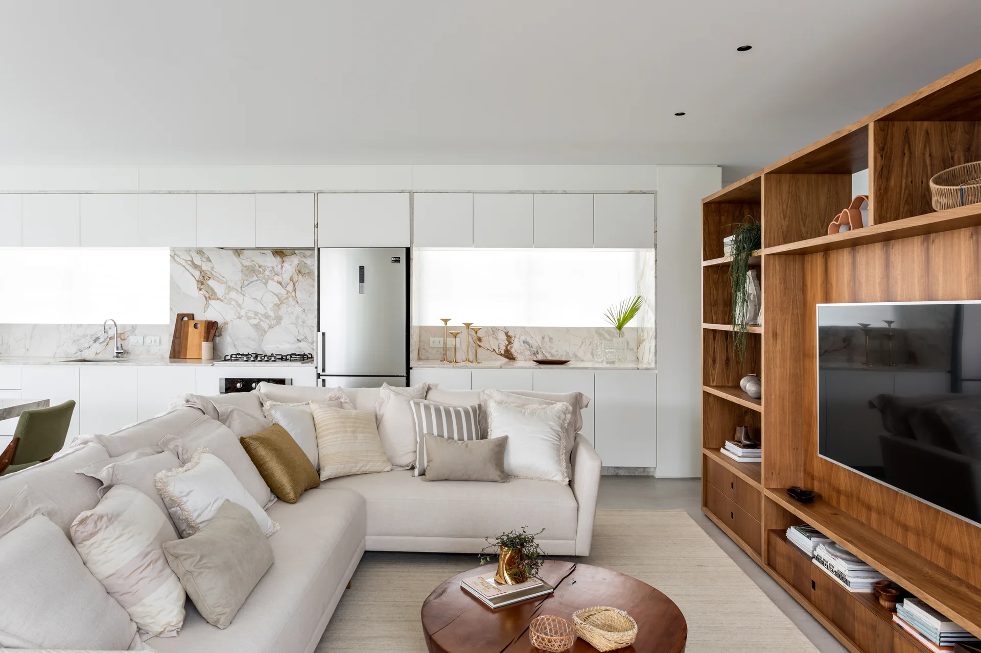 A modern open-plan living room features a cream L-shaped sectional sofa, a round wooden coffee table, and a large wood built-in media shelving unit.