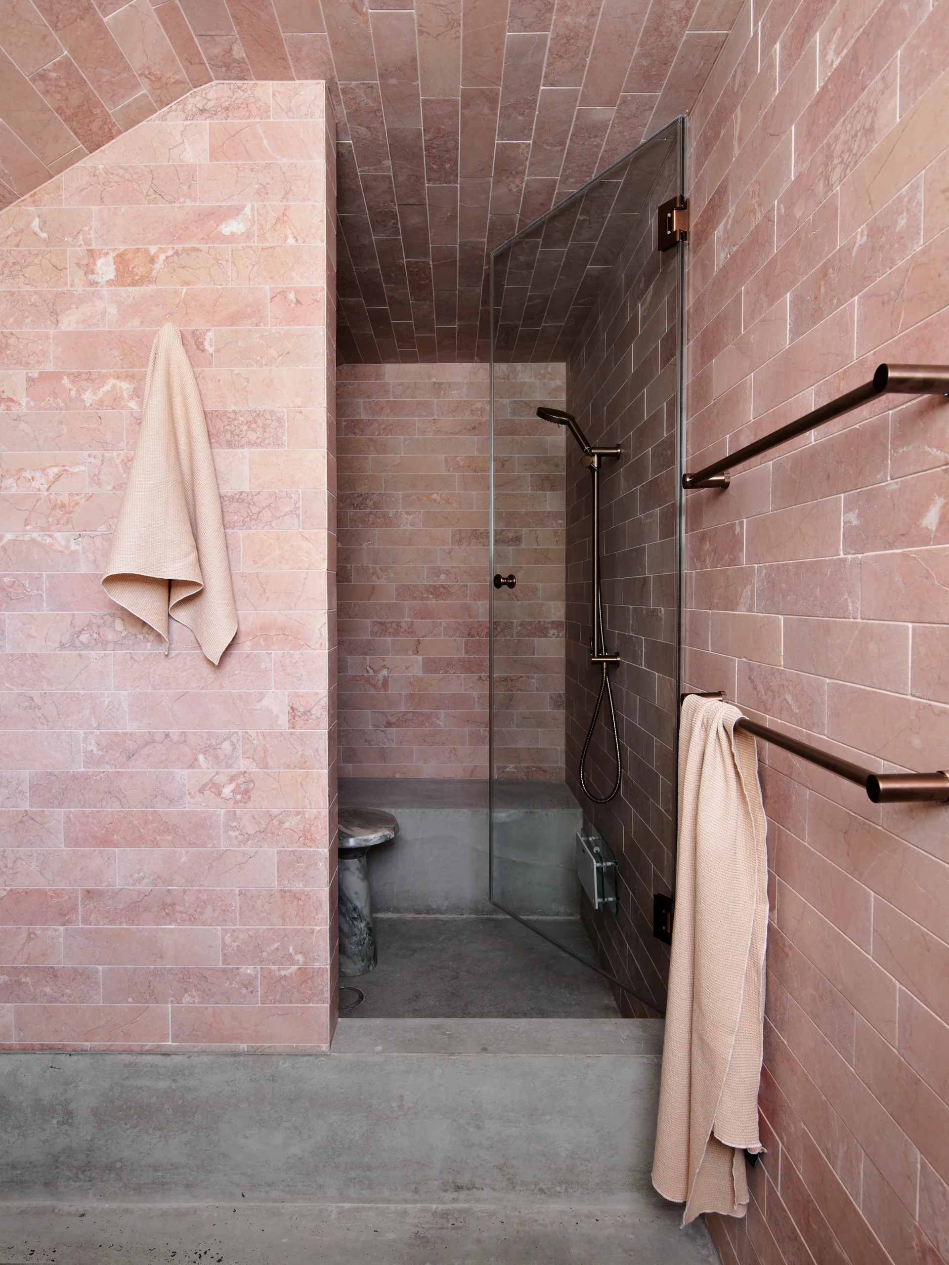 A minimalist bathroom featuring walls and a vaulted ceiling clad in pink stone tiles, a glass shower enclosure, and a concrete floor.