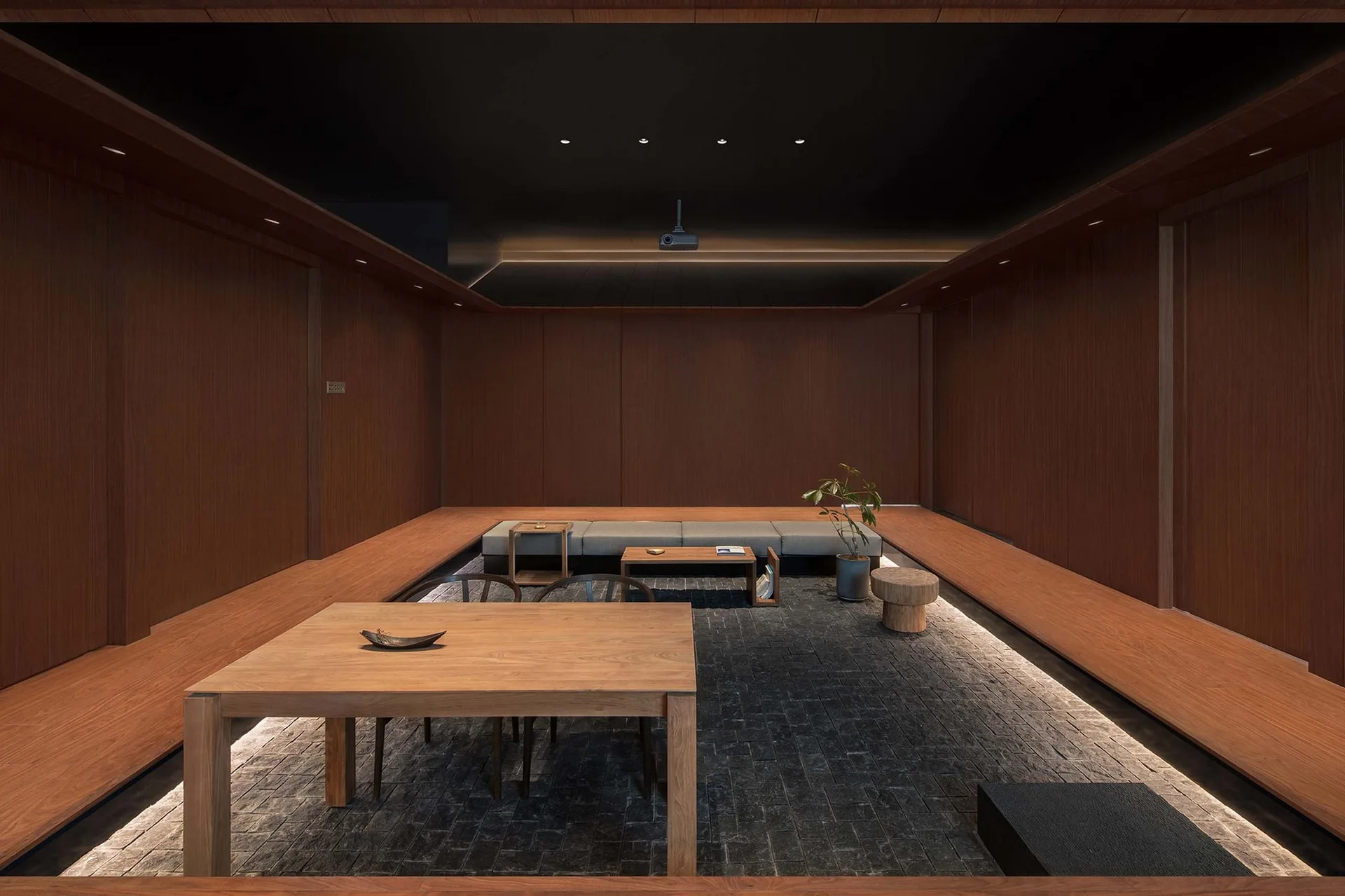 A minimalist sunken living room features warm wood-paneled walls, a low wooden dining table, and a grey tile floor under a black ceiling.