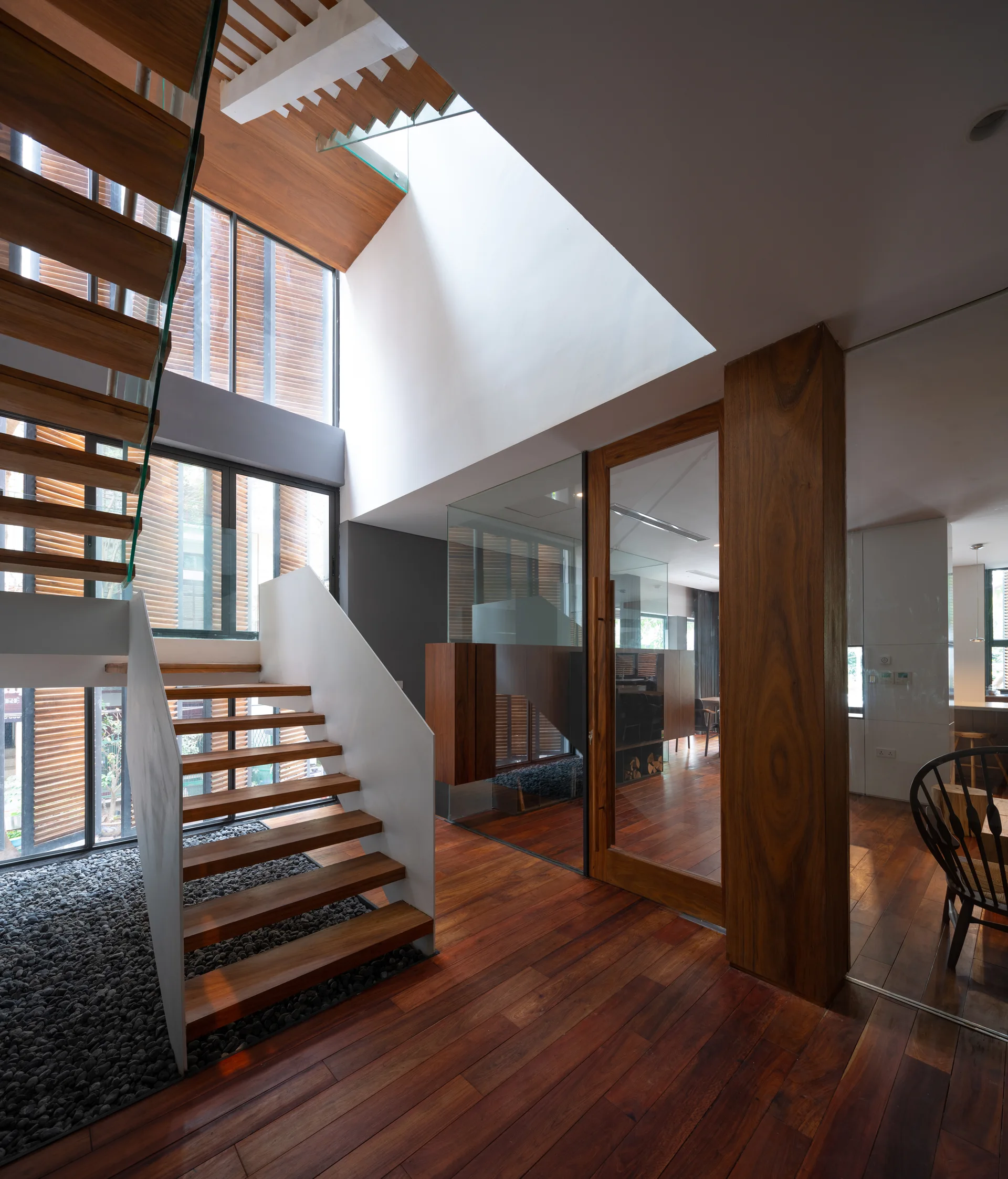 A modern entryway featuring a floating wooden staircase with open treads over a bed of grey pebbles, next to large windows with wood louvers.