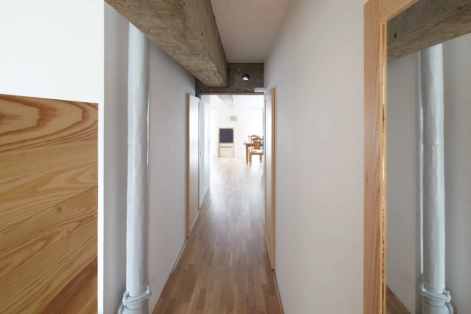A minimalist hallway with light wood flooring, clean white walls, a prominent exposed concrete ceiling beam, and a vertical painted pipe.