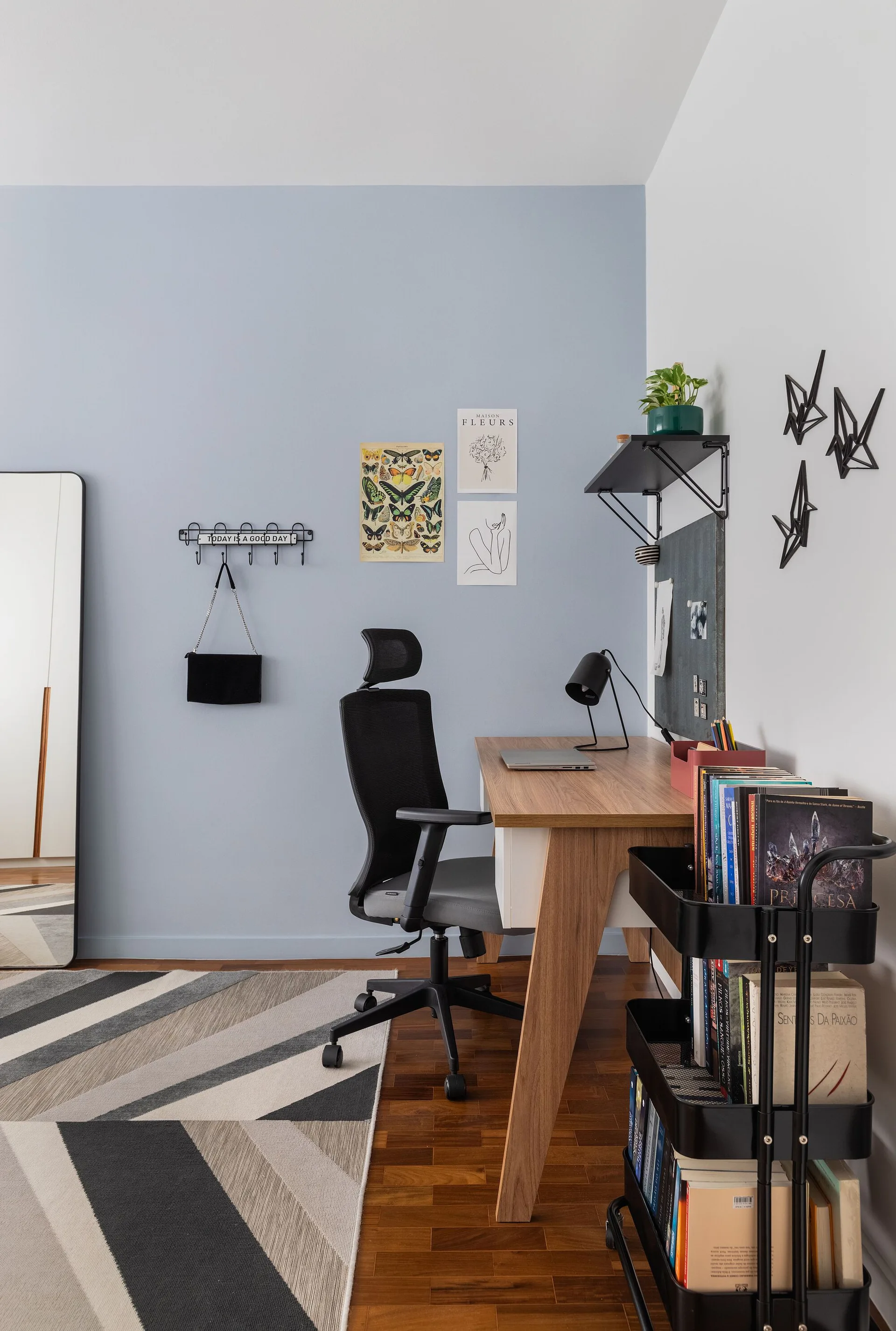A home office workspace features a wooden desk, black ergonomic chair, light blue accent wall, and a black rolling storage cart stacked with books.
