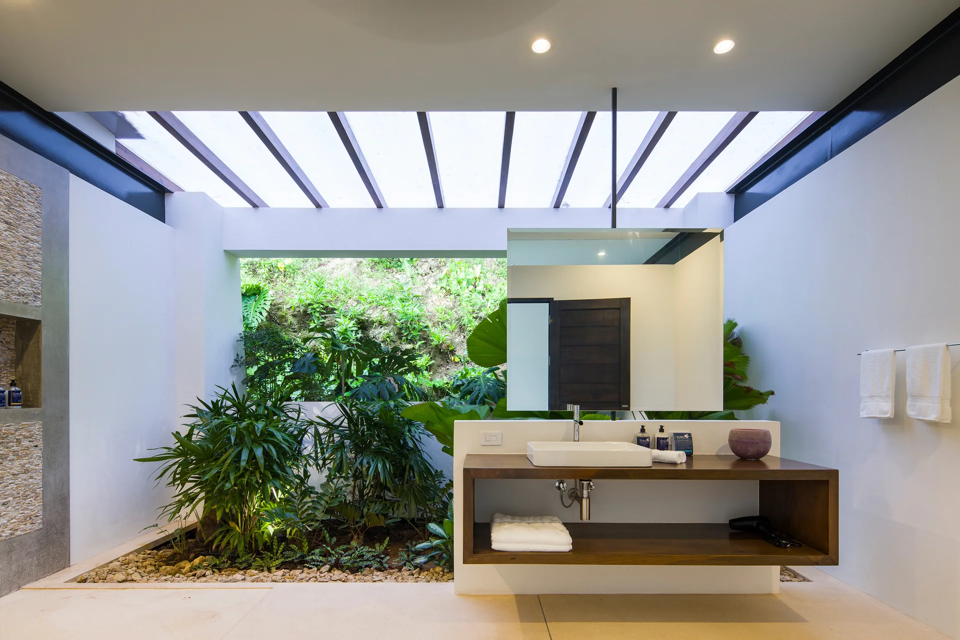 A modern bathroom features a floating wooden vanity with a vessel sink under a suspended mirror, adjacent to an indoor garden beneath a skylight.