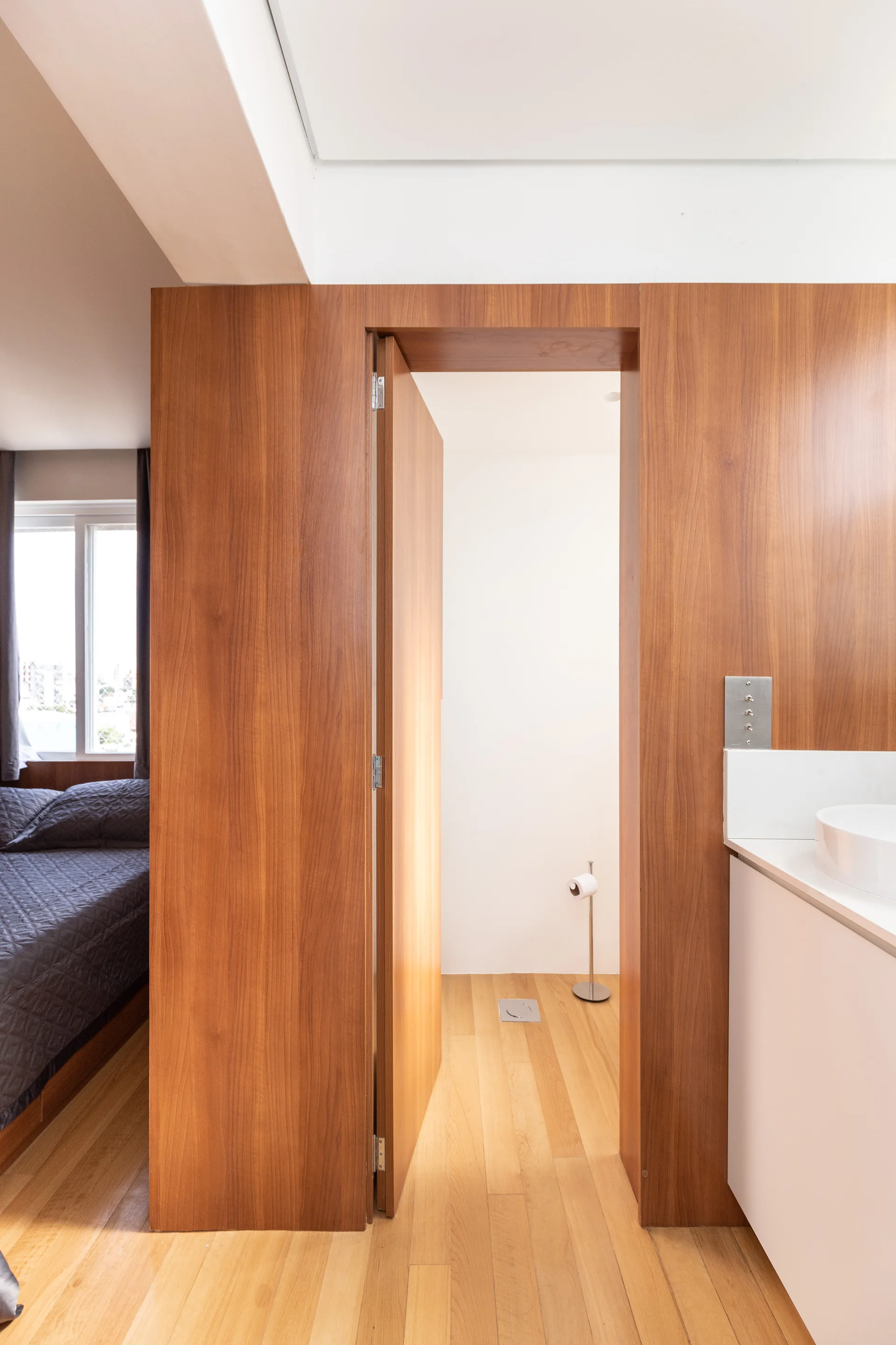 A warm wood-paneled wall with an open doorway connects a bedroom to a bathroom featuring a white vanity with a round sink.