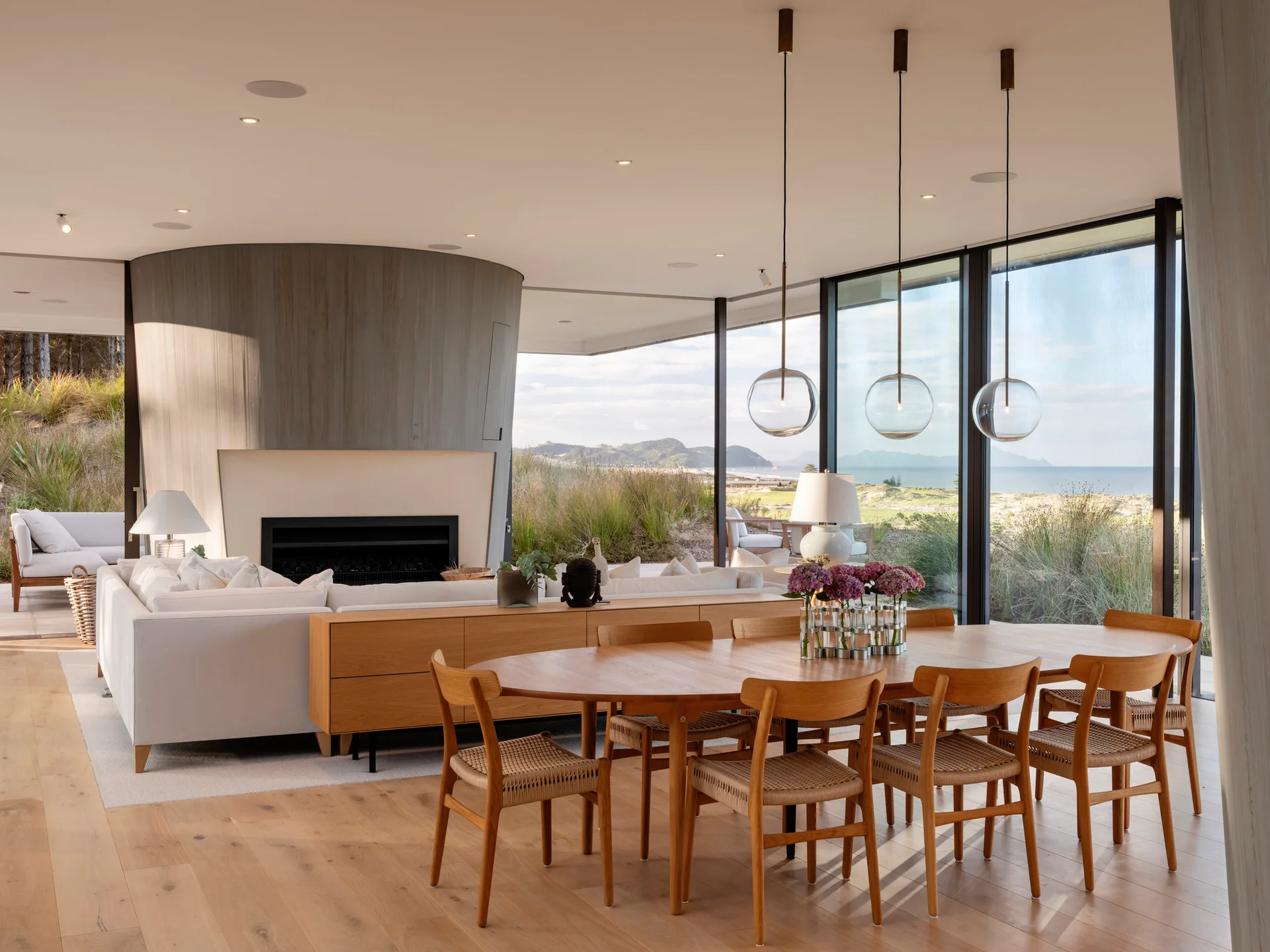 An open dining area features an oval wood table and woven chairs beneath globe pendant lights, with large windows looking out to a coastal landscape.