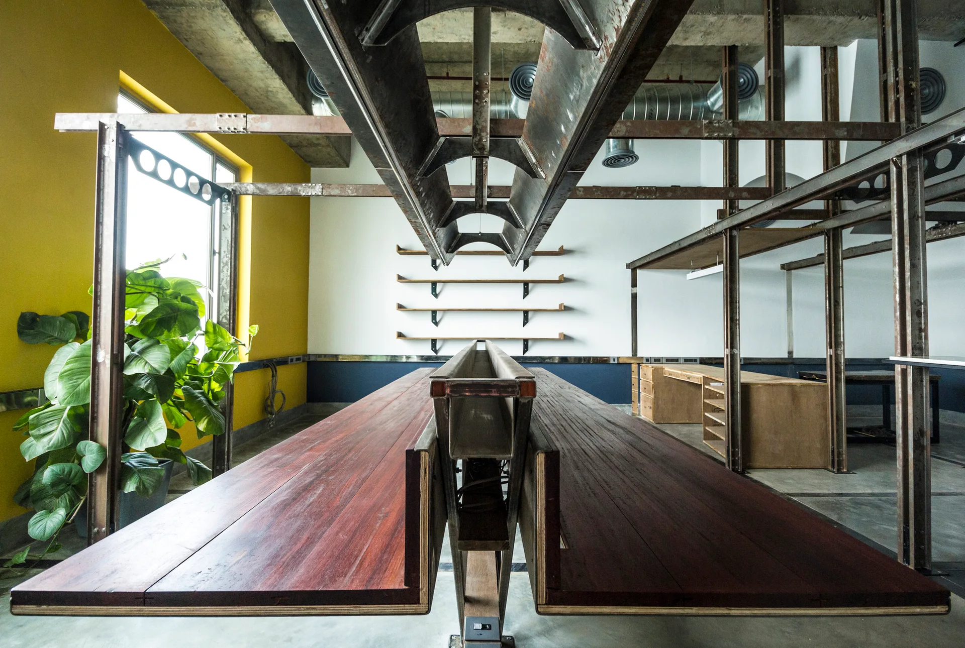 An industrial office workspace features a long central wooden desk beneath exposed steel beams, with a bright yellow accent wall and floating shelves.