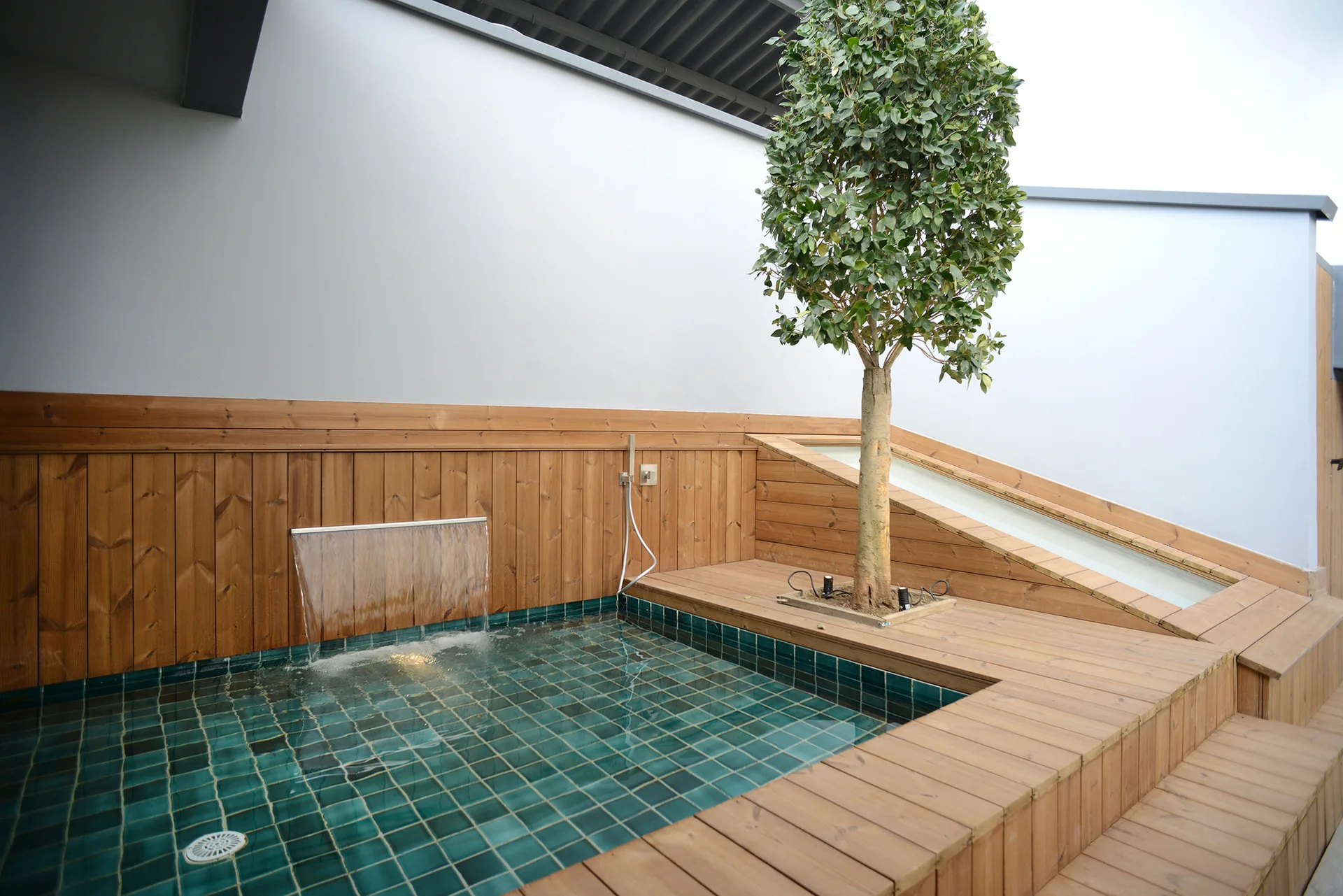 A small outdoor plunge pool with teal tiles and a wall-mounted waterfall is integrated into a raised wood deck with a potted tree.