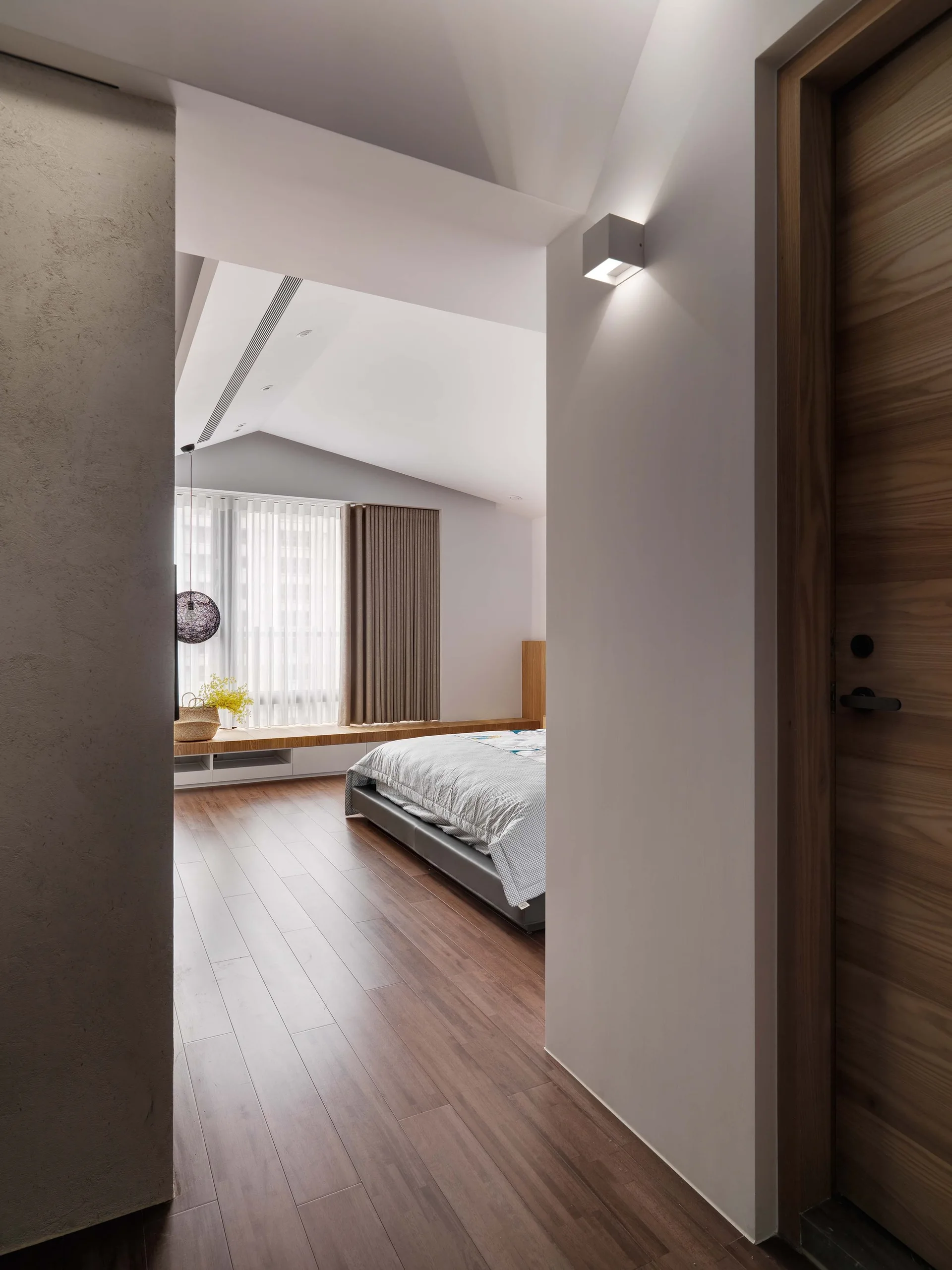 A view from a hallway into a minimalist bedroom with a sloped ceiling, low-profile bed, and a long built-in wooden window bench.