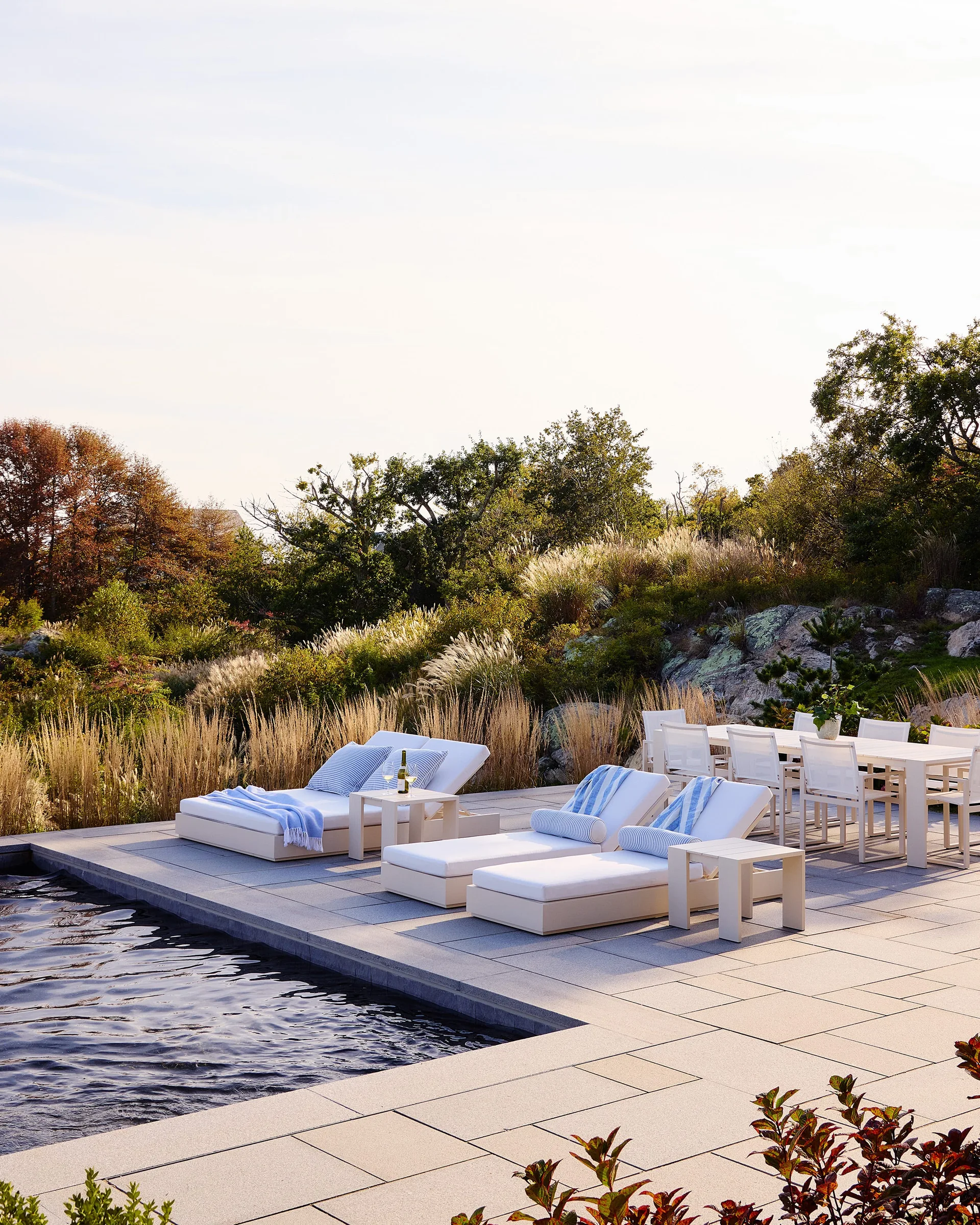 White chaise lounges with blue-striped accents and a matching outdoor dining set sit on a grey stone patio beside a swimming pool.