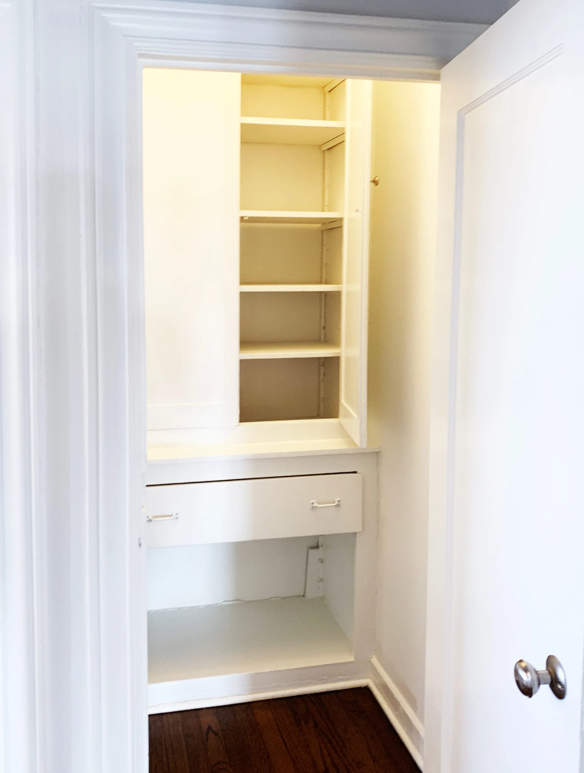 A white built-in storage cabinet with open shelving and a single drawer is recessed within a closet niche featuring dark hardwood floors.