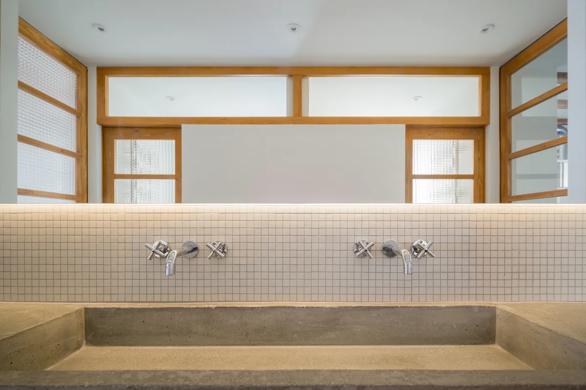 A close-up of a modern bathroom vanity with a grey concrete trough sink, wall-mounted faucets, and a white mosaic tile backsplash.