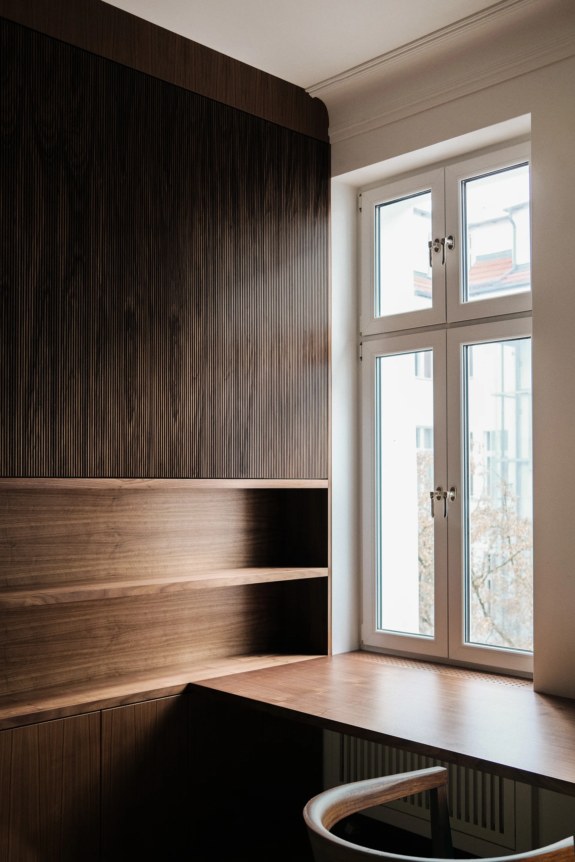 A custom walnut built-in desk and shelving unit with fluted upper cabinets sits next to a tall white window with an integrated slatted radiator cover.
